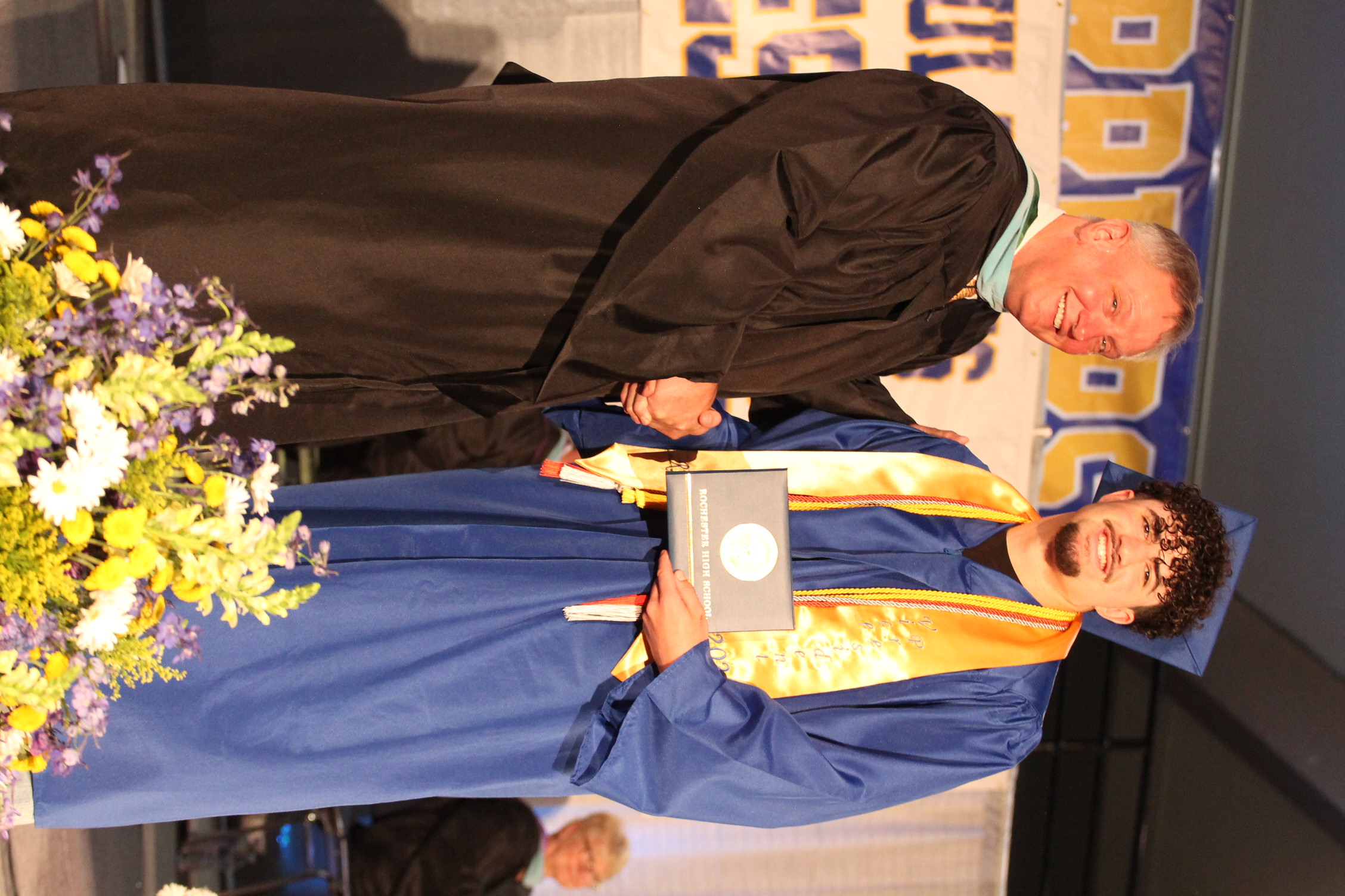 Two men in graduation attire stand on a stage, one shaking hands with another holding a certificate.