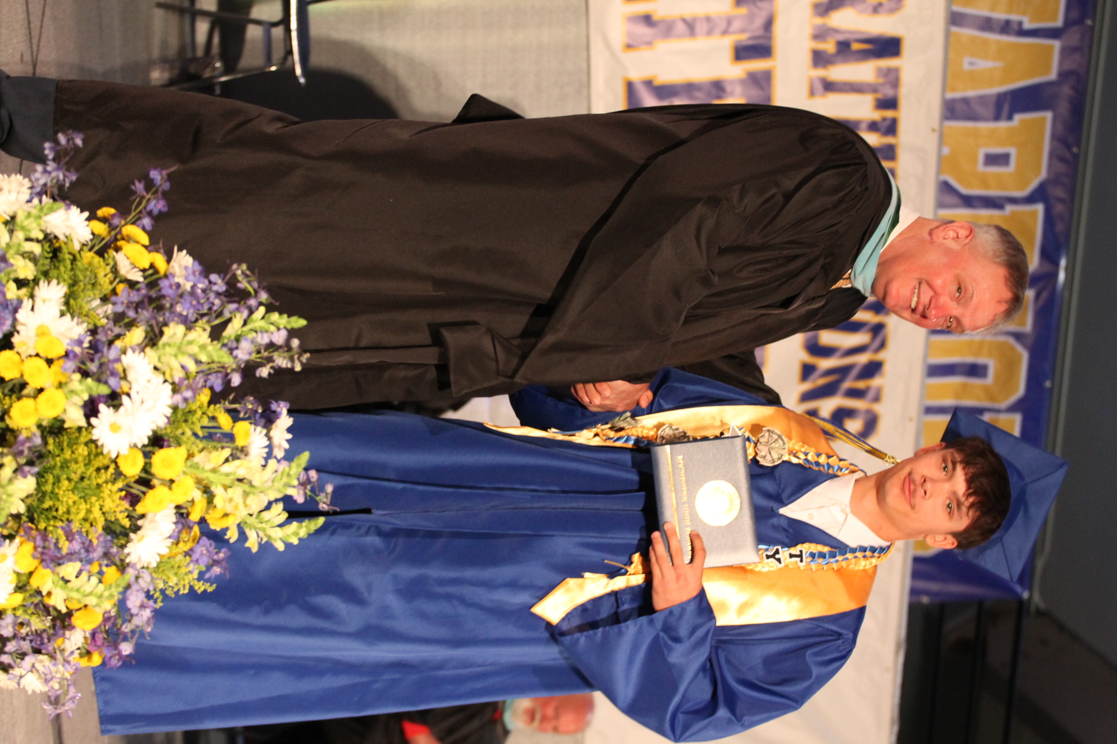 A male student in a blue gown and cap receives an award from a faculty member.