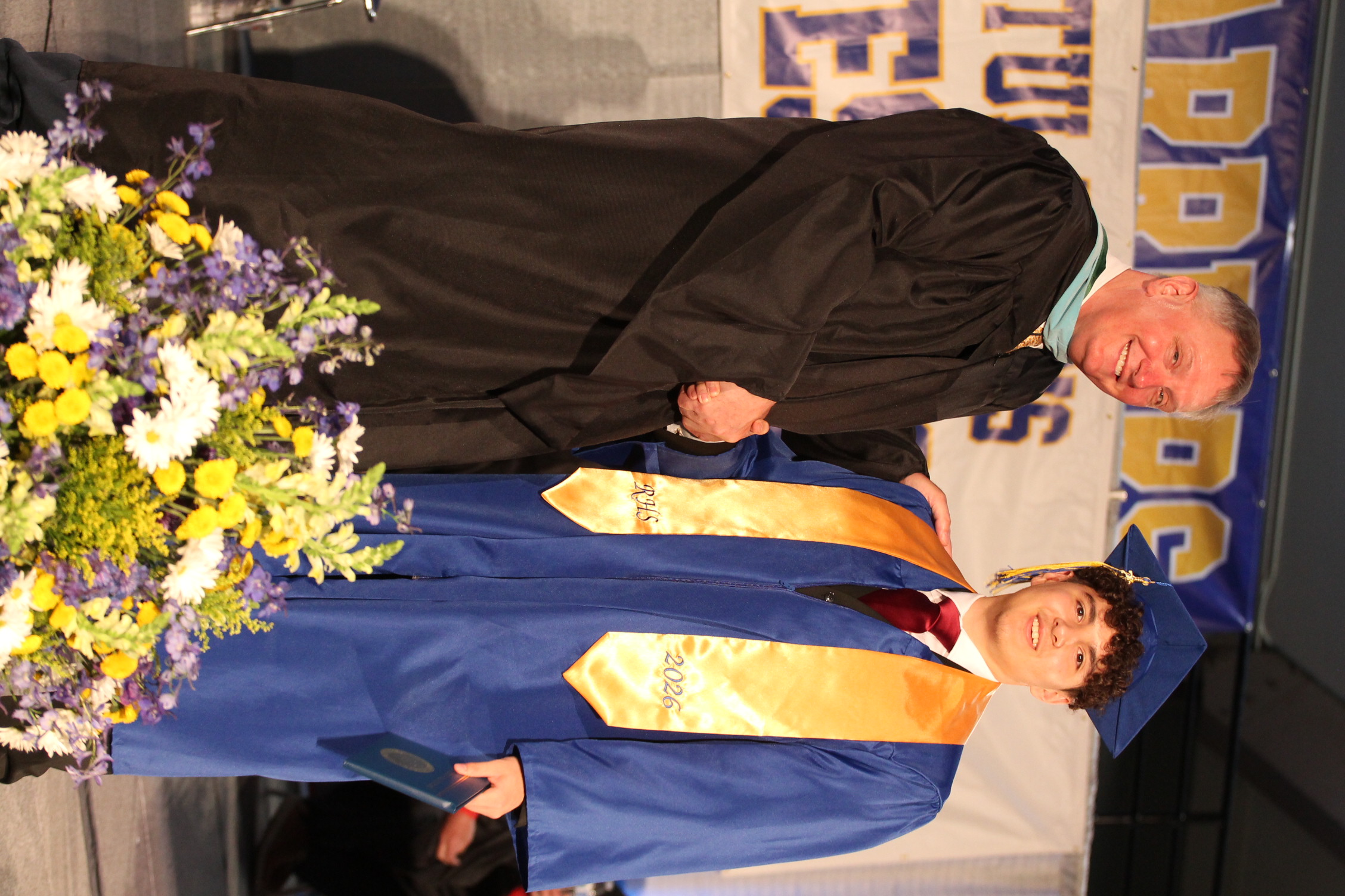 A man in a black gown, likely a faculty member, shakes hands with a graduate wearing a blue gown and yellow sash.