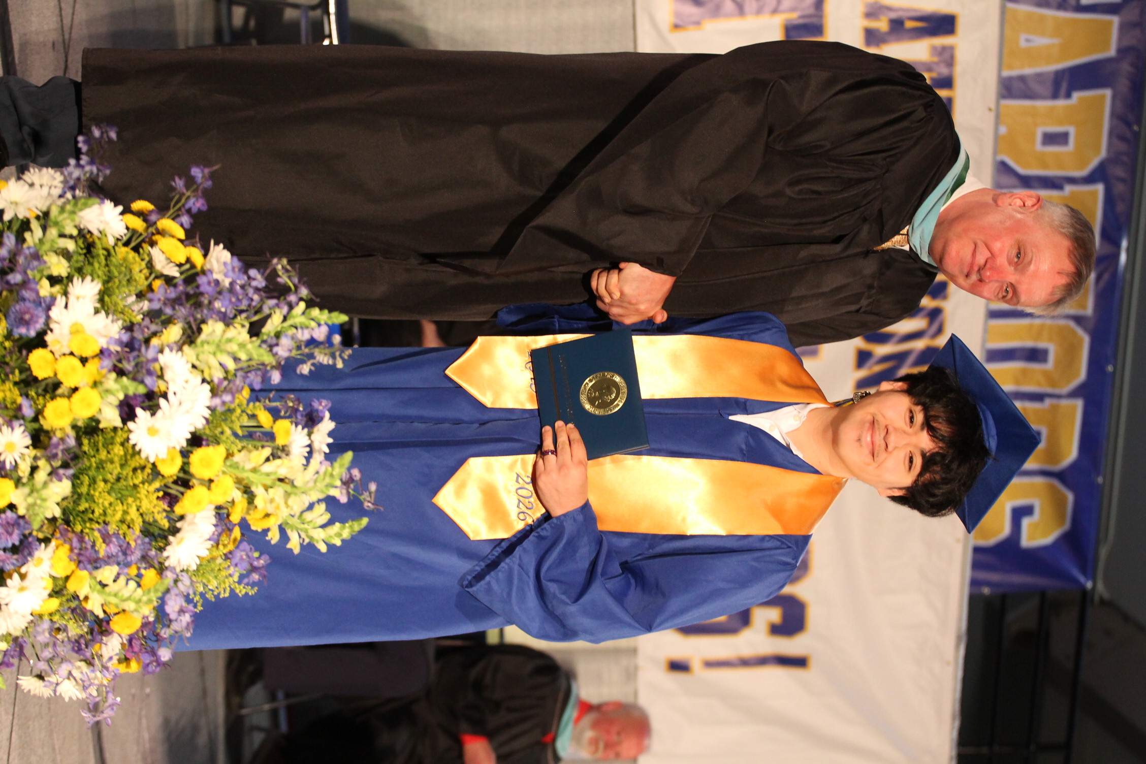 A man in a black gown hands a diploma to a young woman in a blue gown.