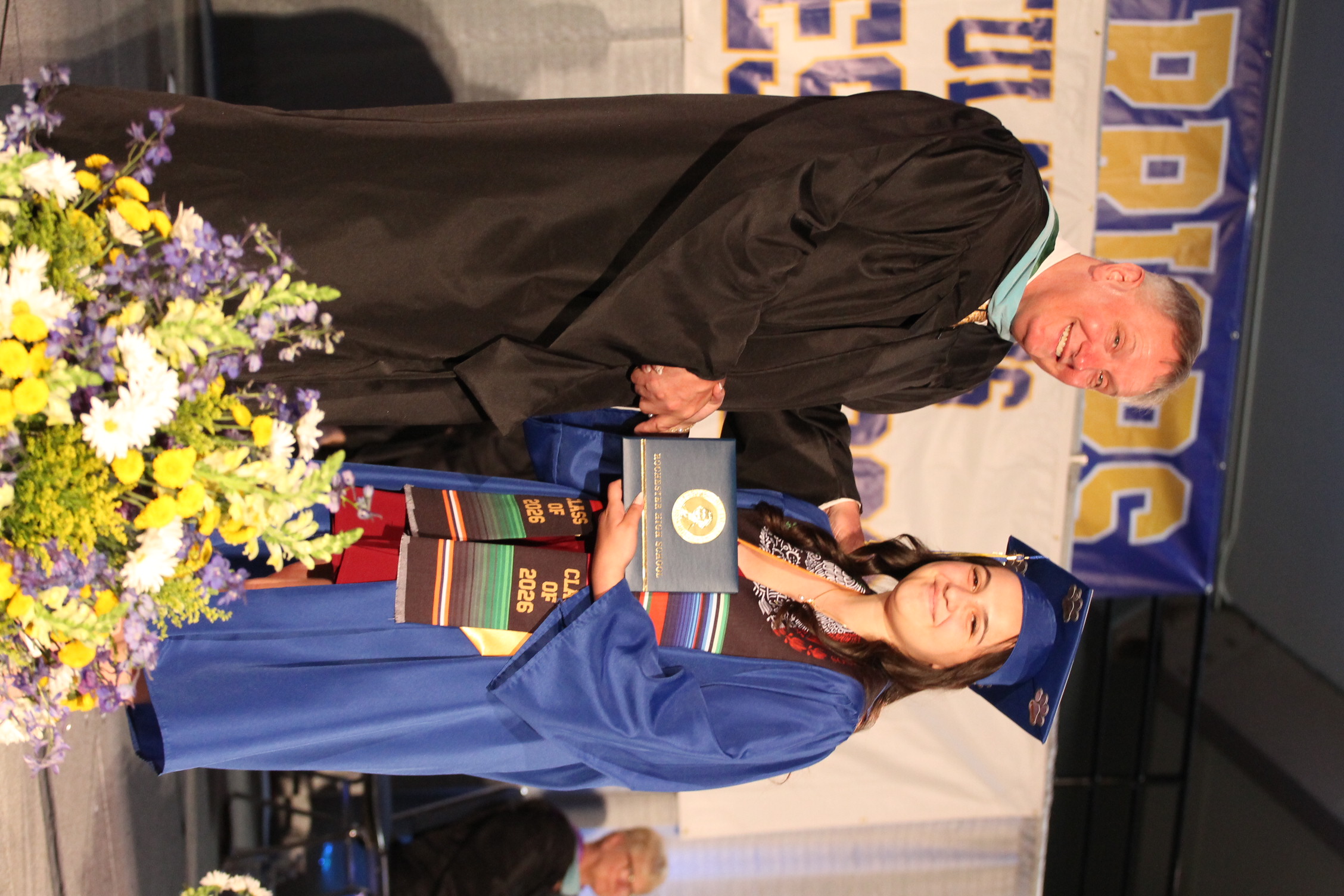 Man in black robe shakes hands with a woman in blue gown. She holds an award plaque. Floral arrangement on stage.