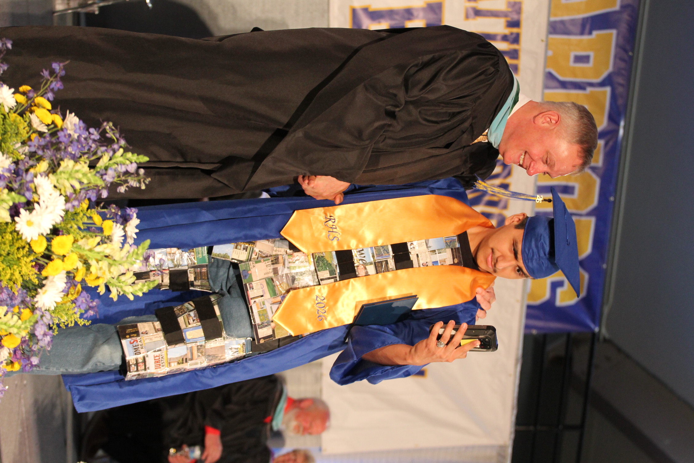 A man in a graduation gown is taking a selfie on stage, accompanied by an older man in a cap and gown.