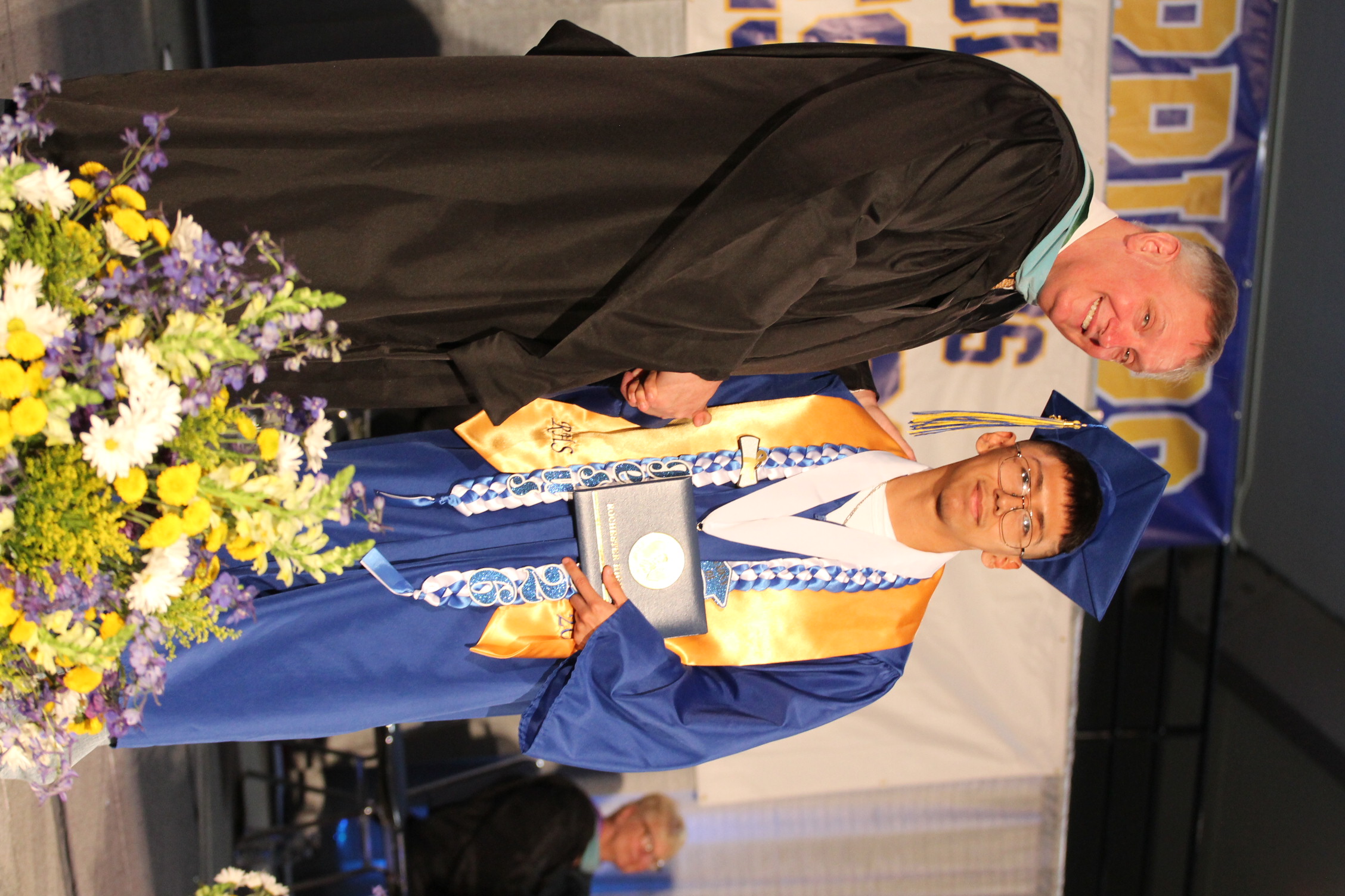 A man in a black gown presents an award to a student in a blue gown. Both stand on a stage with flowers.
