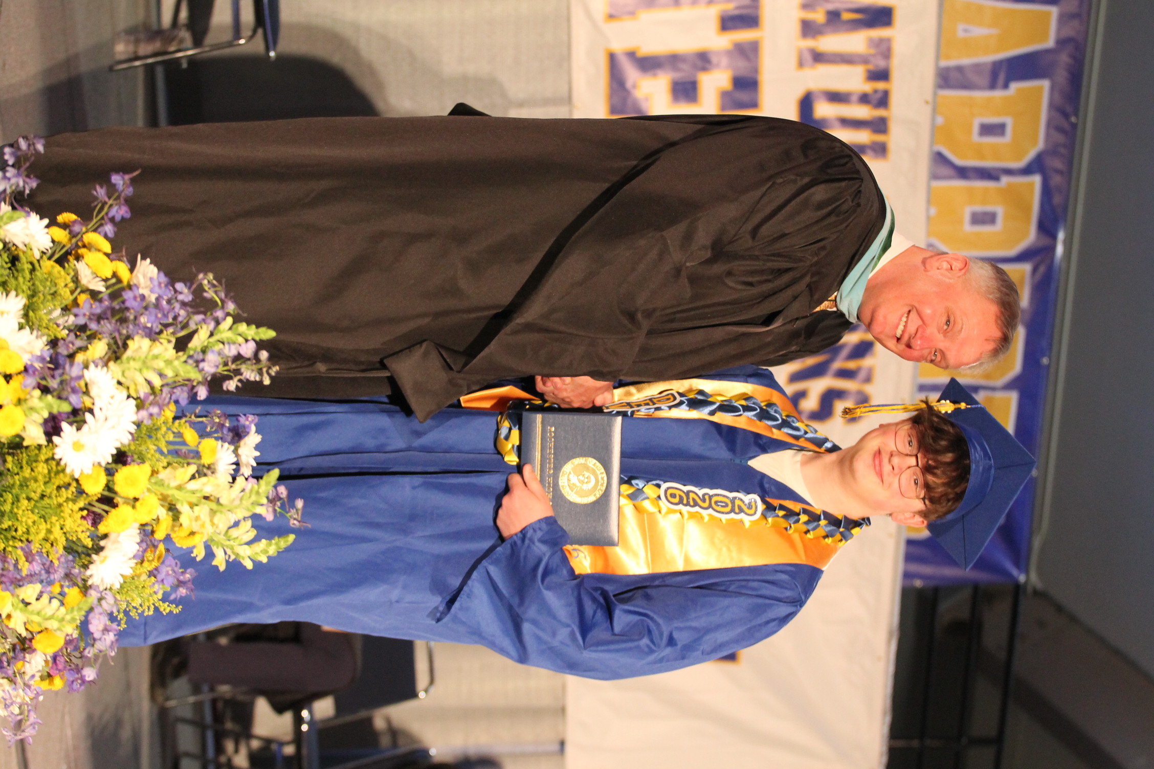 A man in a black robe stands beside a graduate in blue, holding a diploma, with flowers in front.