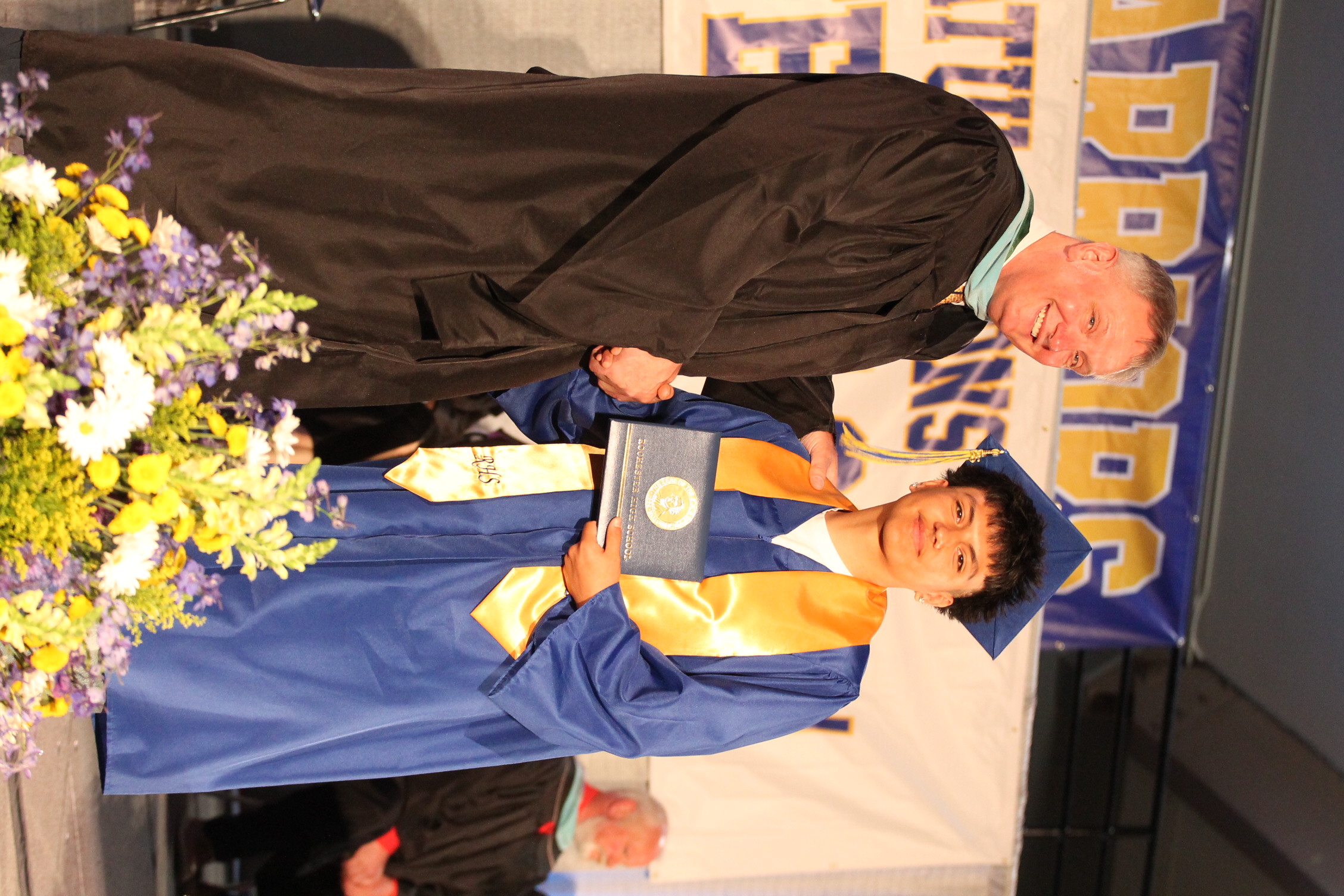 A man in a black robe shakes hands with a graduate in a blue gown, who holds a certificate. Behind them is a stage with a flower arrangement and a banner.