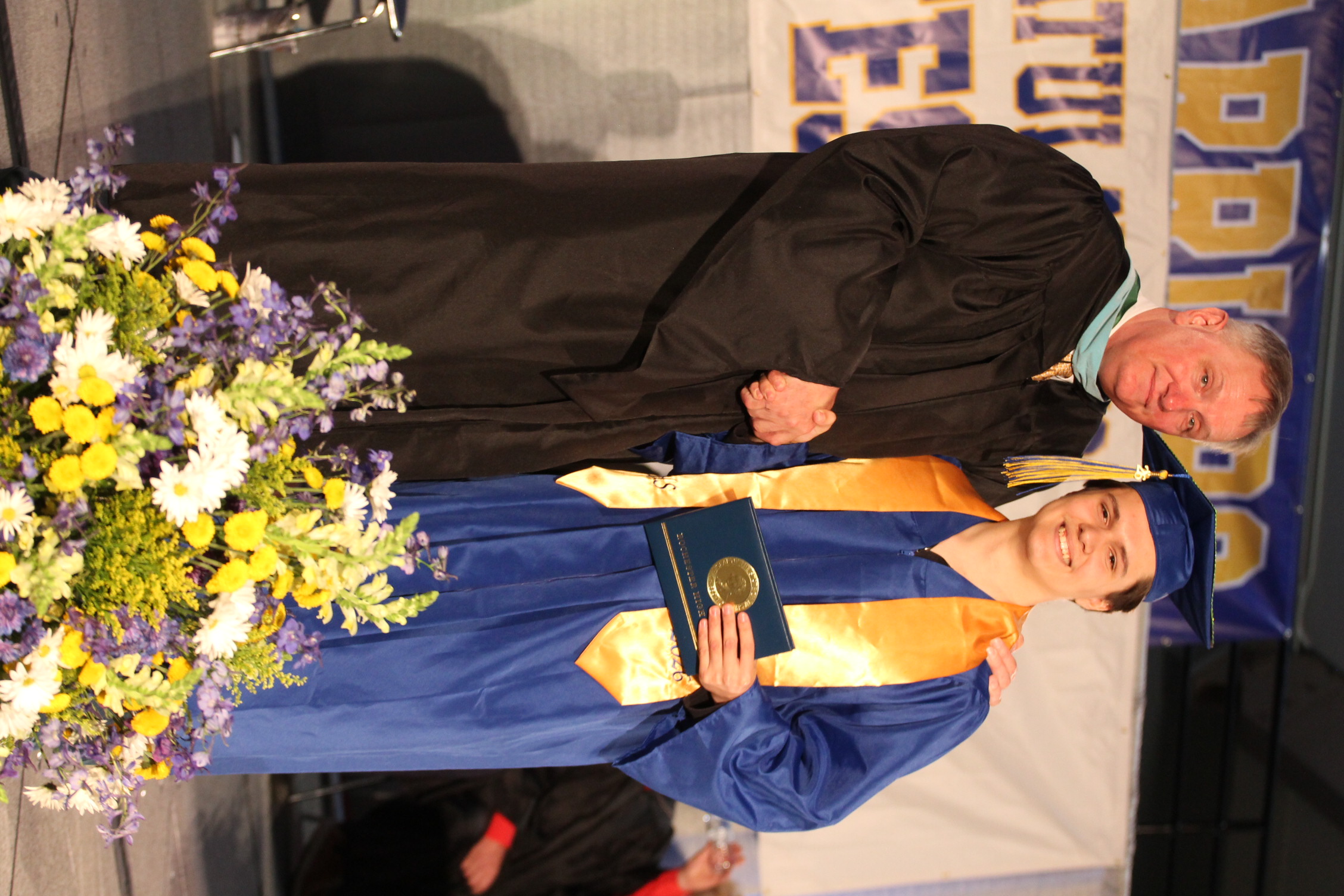 A man and a woman, both dressed in graduation gowns, are standing next to each other on stage.