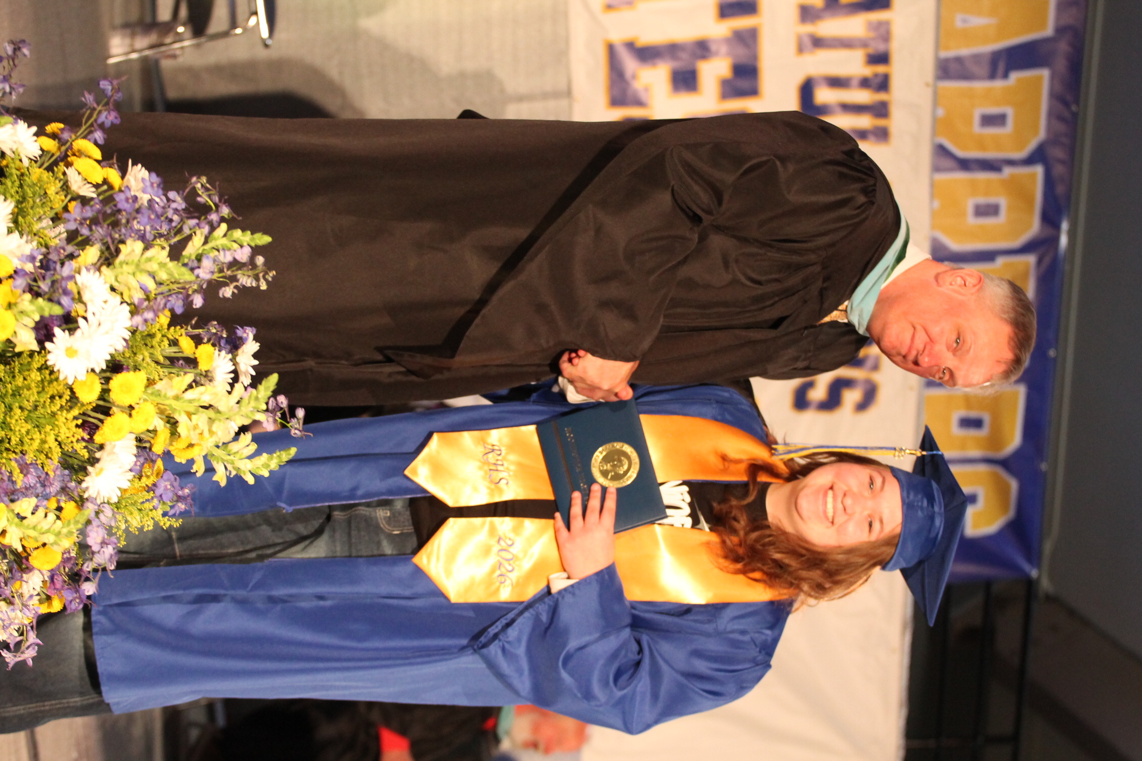 A person in a black gown gives a diploma to a graduate in a blue gown. They are standing in front of flowers.