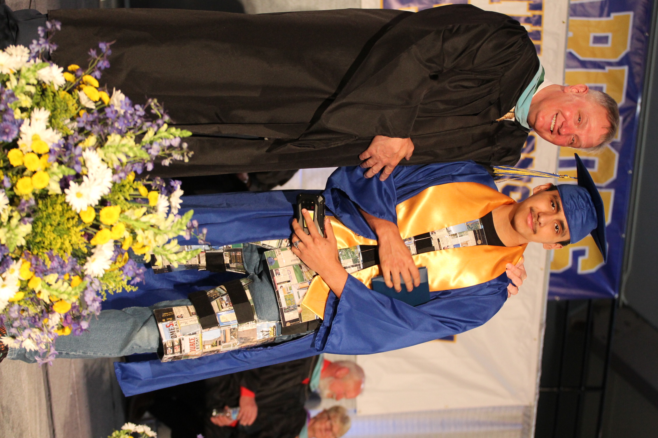 Two men on a stage at an event. One in a black gown, the other in blue, with a flower arrangement nearby.