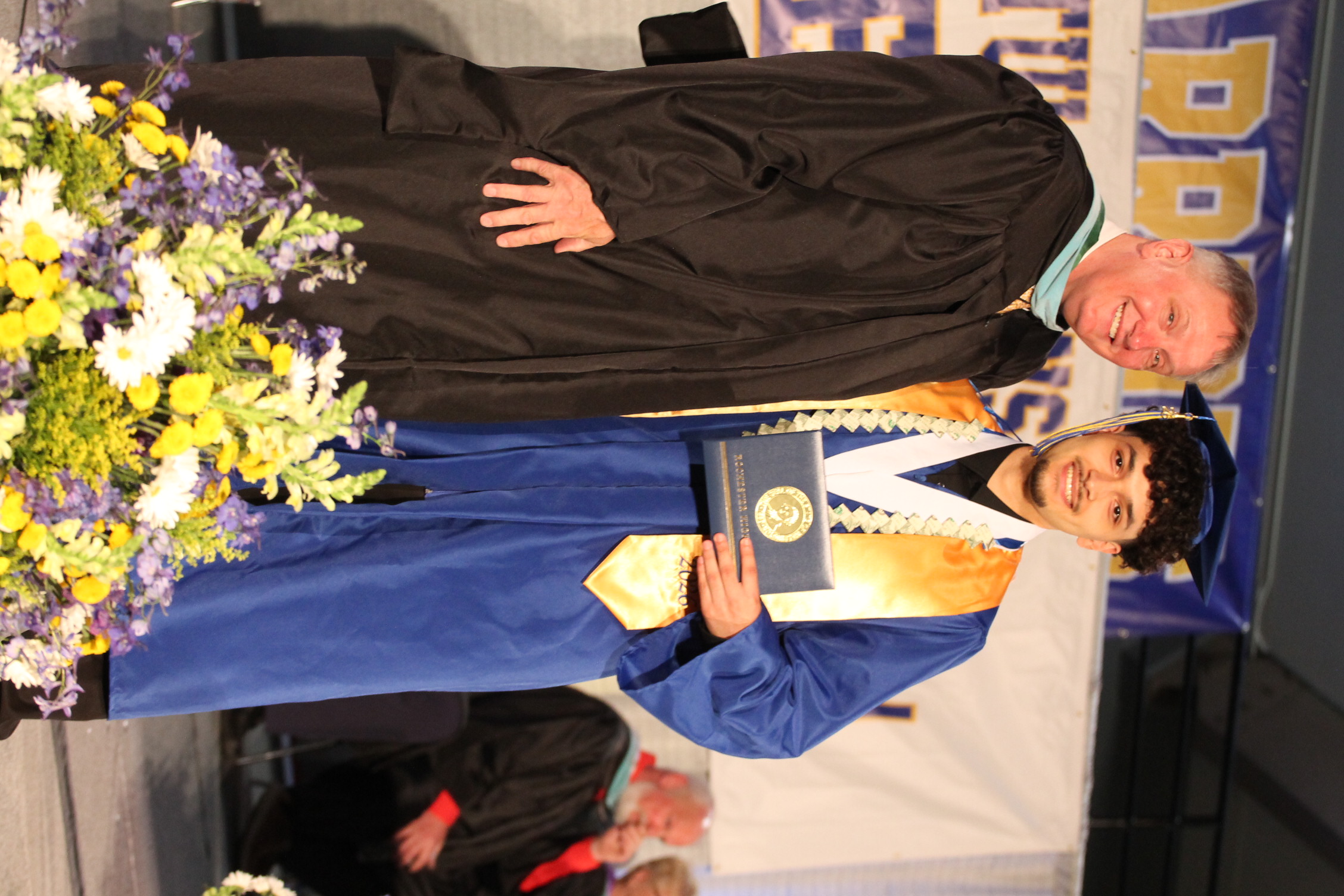 Two men in graduation gowns, one holding an award. The man in blue stands on a stage with flowers.