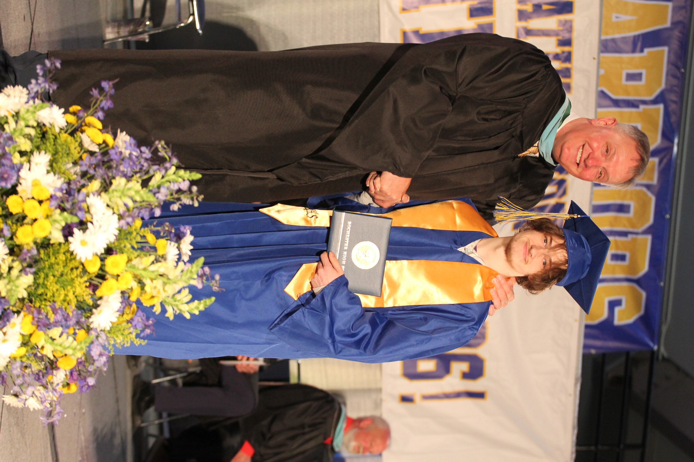 A man and a graduate in academic regalia stand together on stage with flowers. The man holds an award, and a banner is behind them.