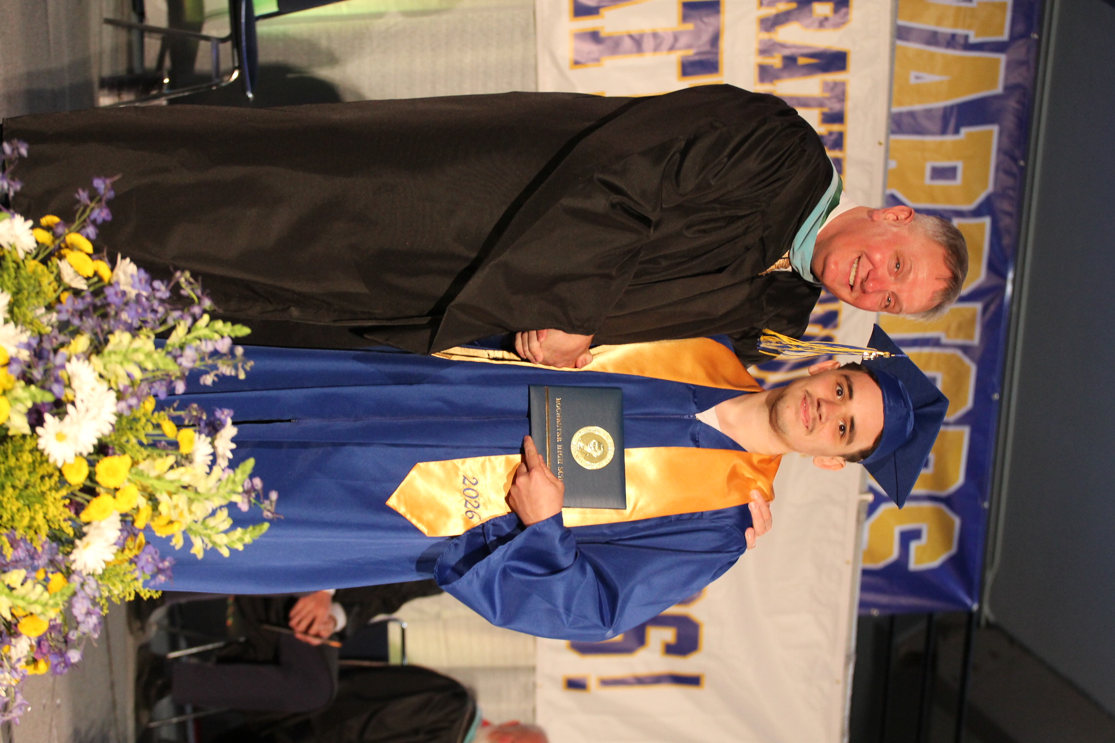 A man in a blue gown holds a diploma, standing next to another man in a black gown, both smiling, in front of a stage with flowers and a banner.
