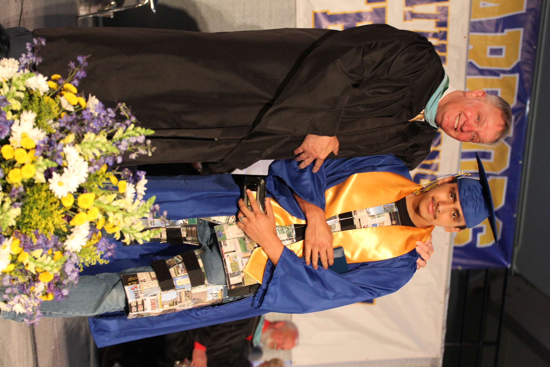 Man in a black gown and a man in a blue gown with a mortarboard cap, standing on stage.