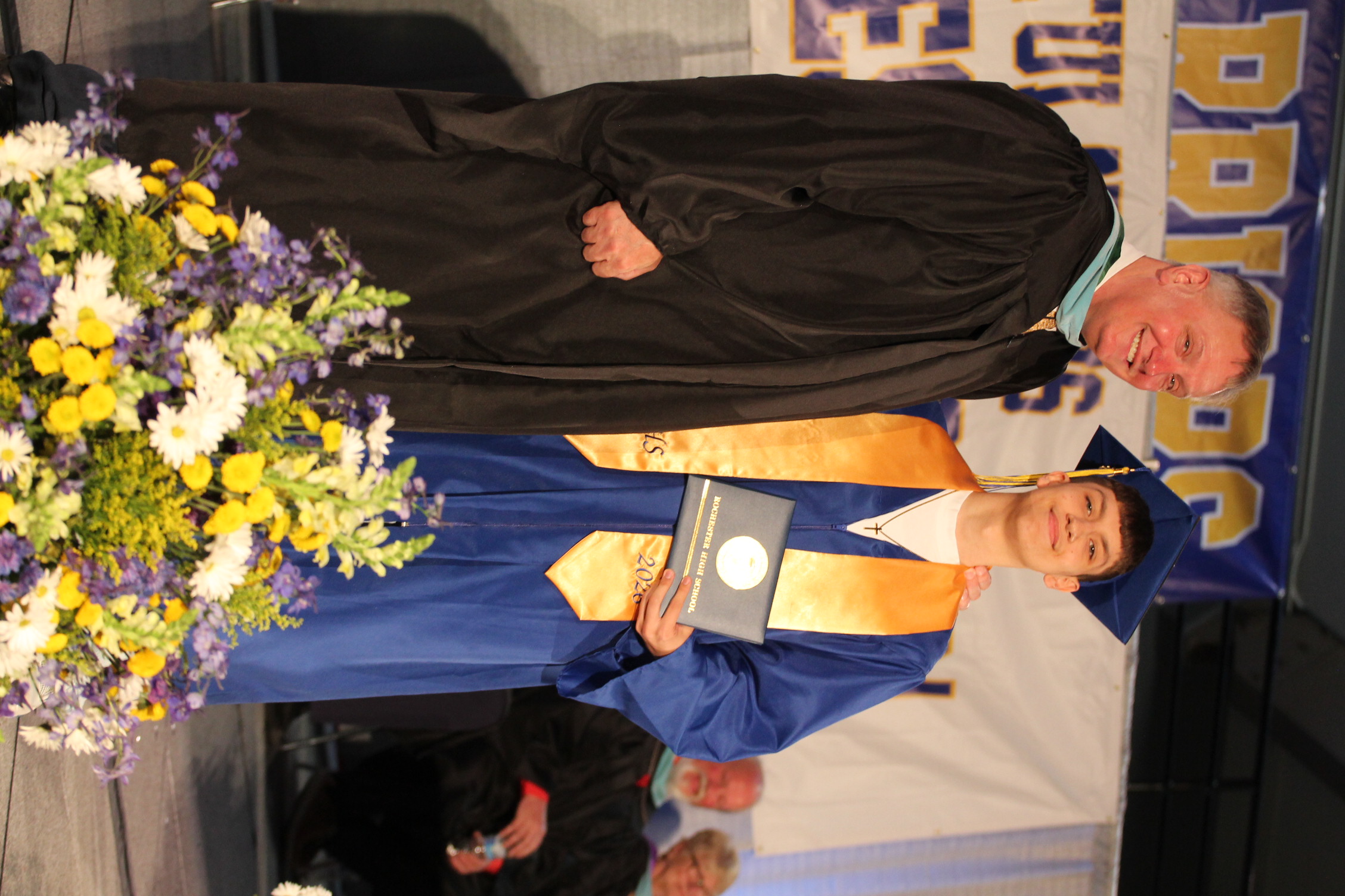A man and a student in graduation robes stand on stage; the student holds a diploma and a bouquet of flowers.