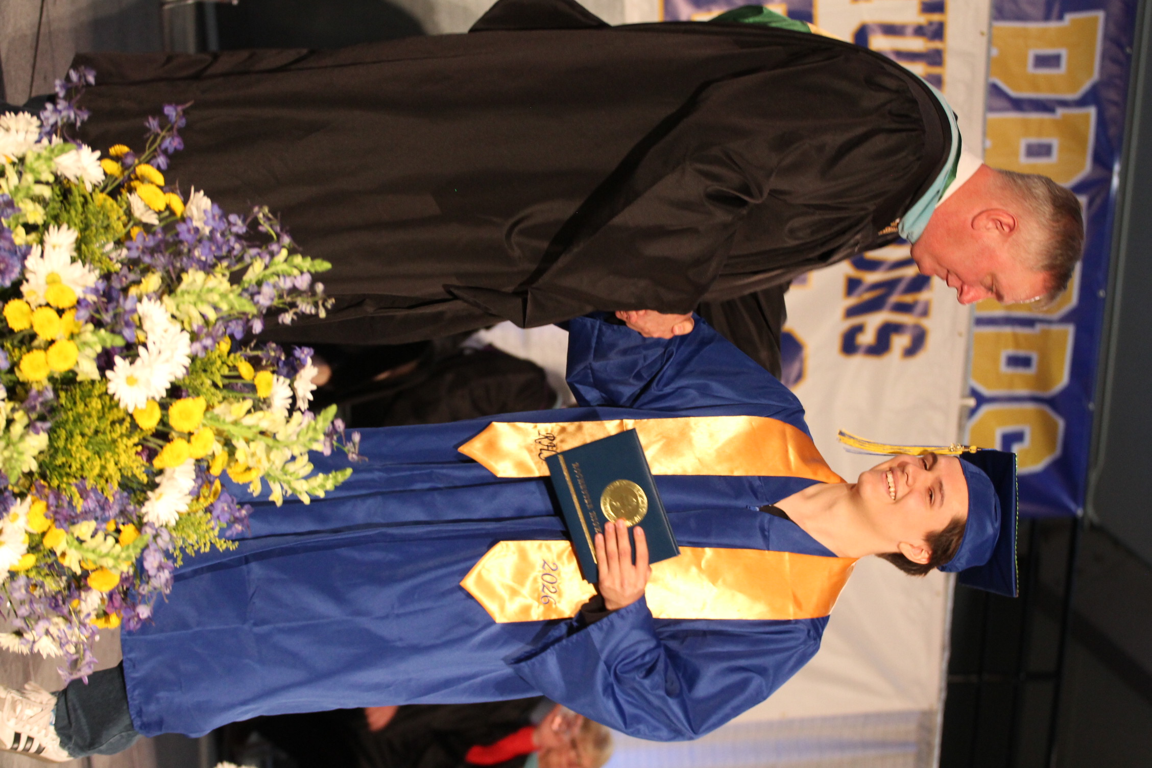 A man in a black gown hands a diploma to a man in a blue gown. They stand on stage next to flowers.