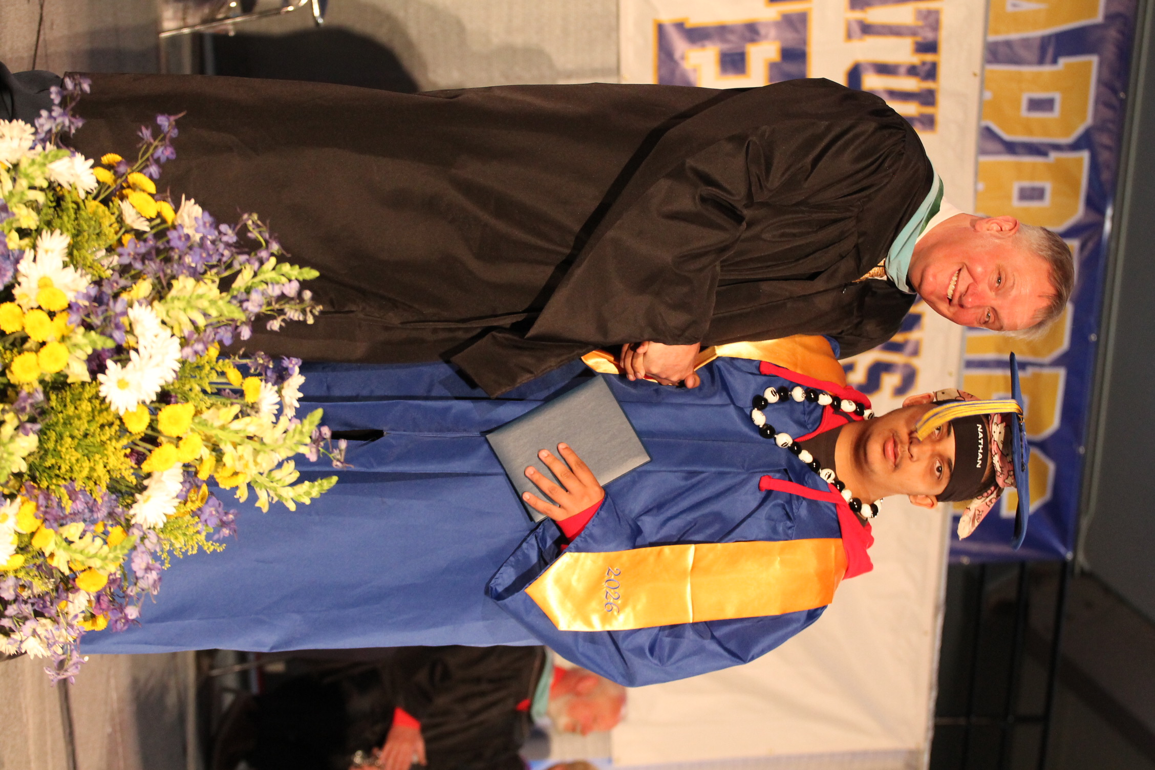 A man in a black gown shakes hands with a graduate in a blue gown, standing on stage.