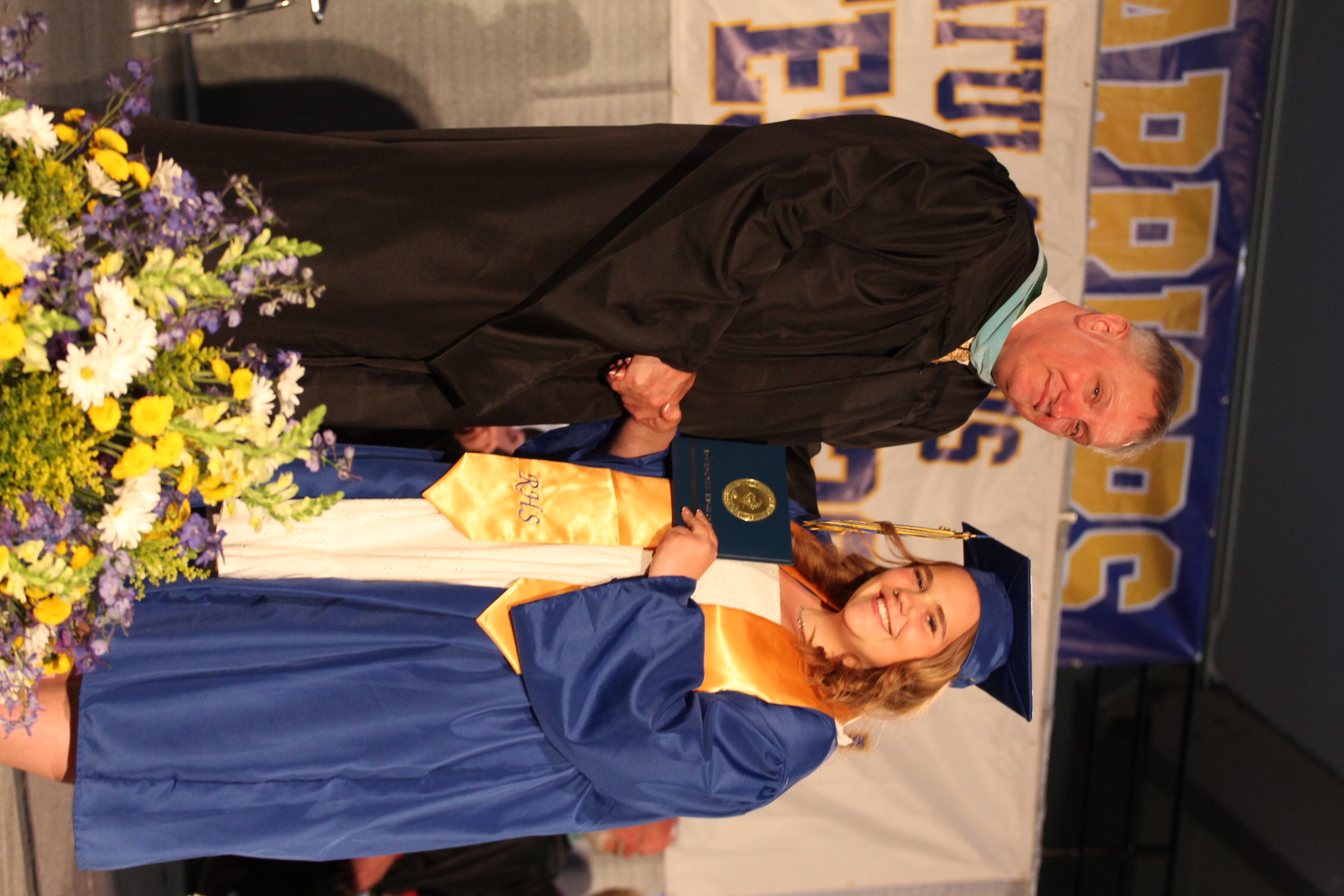 A man and a woman in formal attire, with the man presenting the woman a diploma. The background has a banner with text.