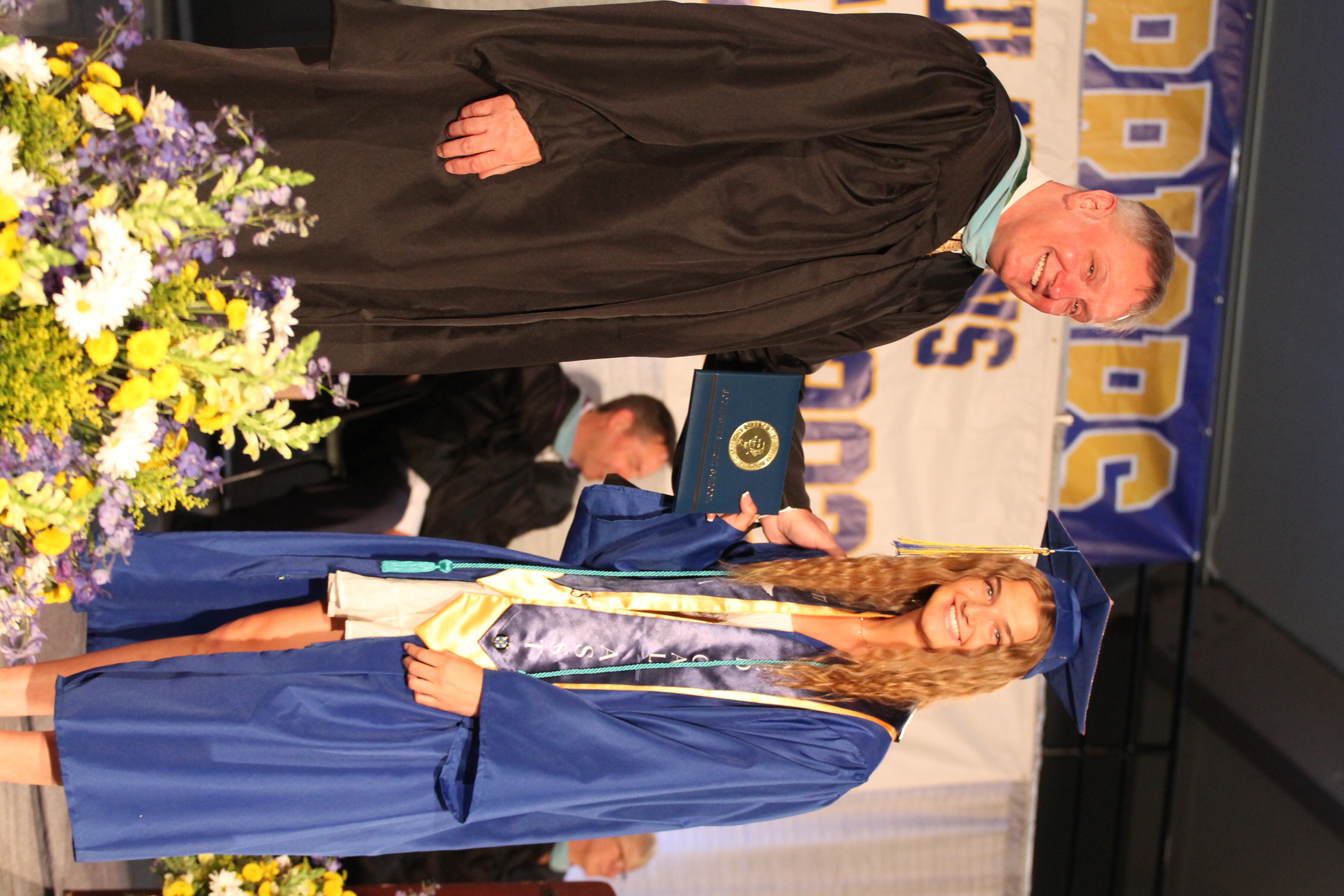 Man and woman on stage. Man in black graduation robe holds a diploma. Woman in blue robe holds diploma.