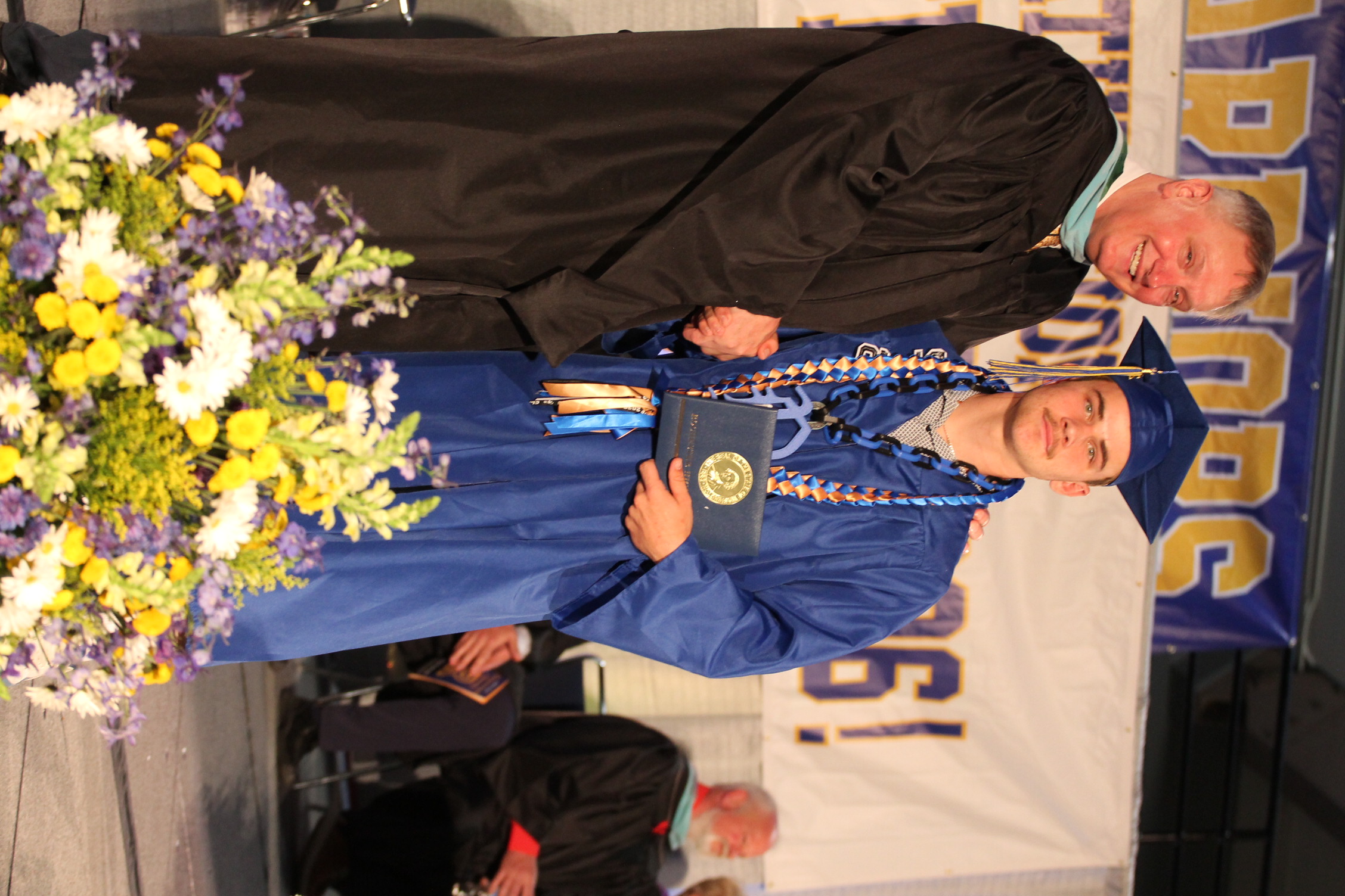 Man in black graduation gown with another man in blue gown and cap holding certificate, standing before flowers.