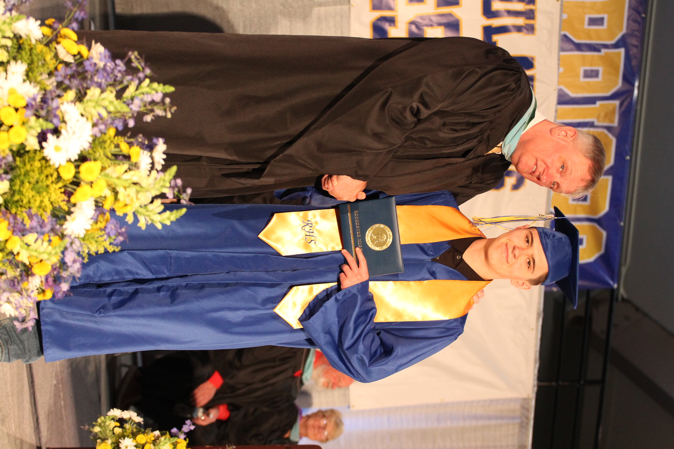 A person in a black gown stands beside a person in a blue gown with a yellow stole, both smiling and holding a diploma.
