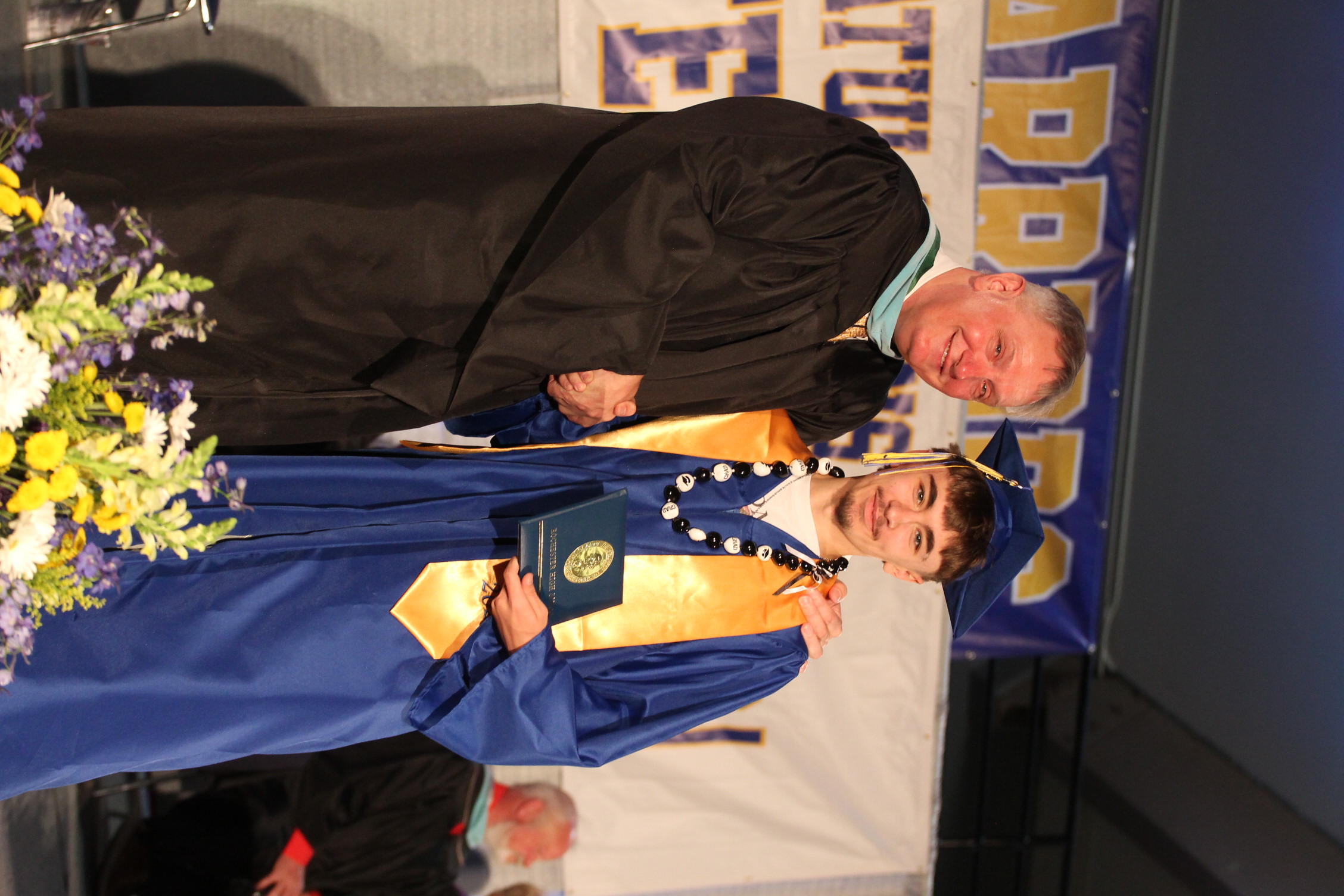 A man in a black gown shakes hands with a graduate wearing a blue gown, both standing on stage.