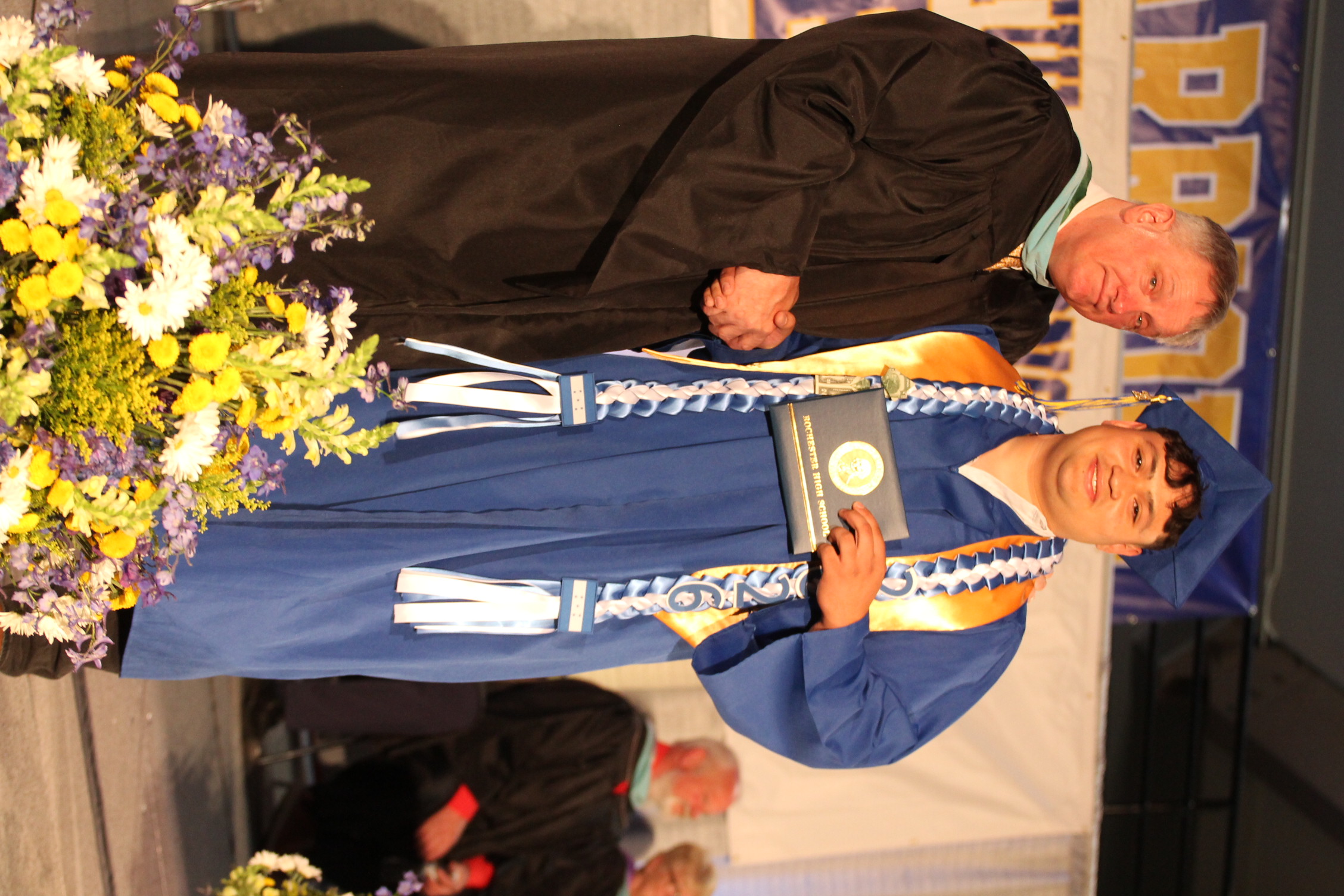 A man in a black graduation gown presents an award to a graduate in a blue gown on a stage with flowers.