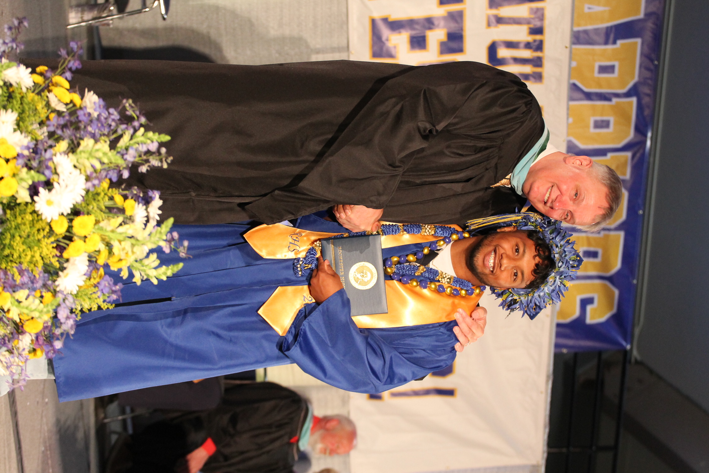 A man in a black graduation gown presents an award to a man in a blue gown.