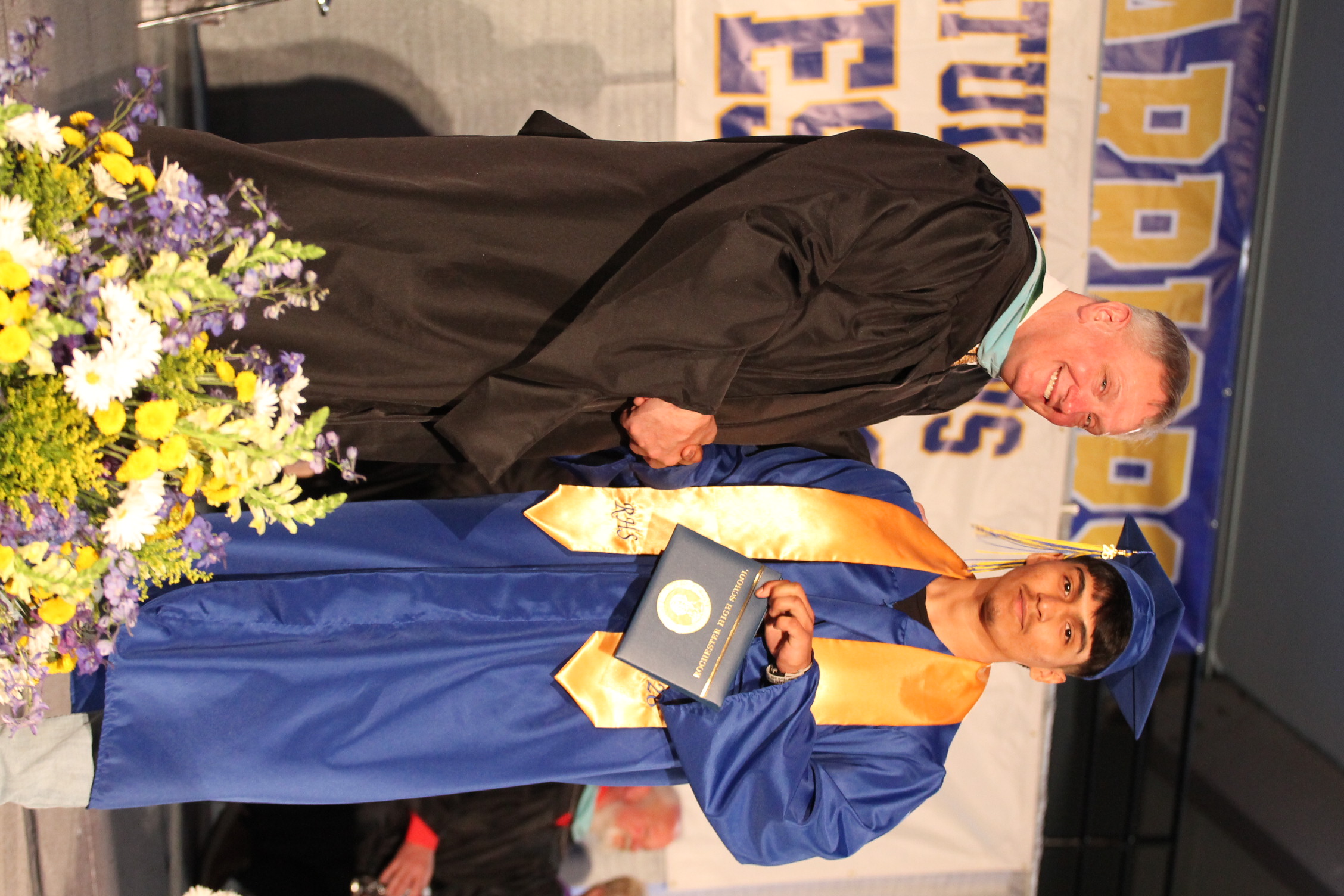 A man in a black gown hands a diploma to a graduate in a blue gown. A floral arrangement is in the foreground.