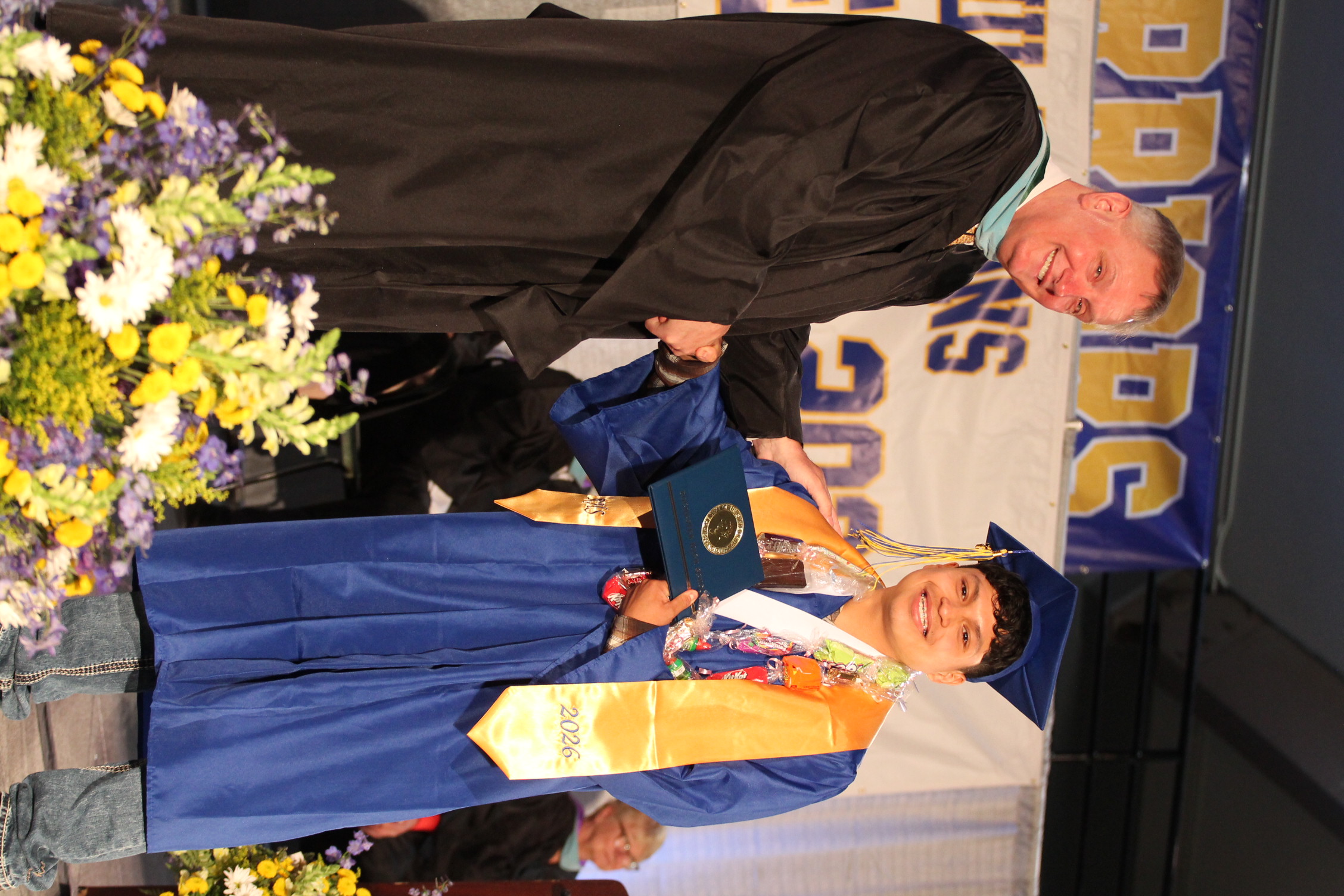 A man in a black gown and another in a blue gown and cap stand before a flower arrangement, shaking hands.