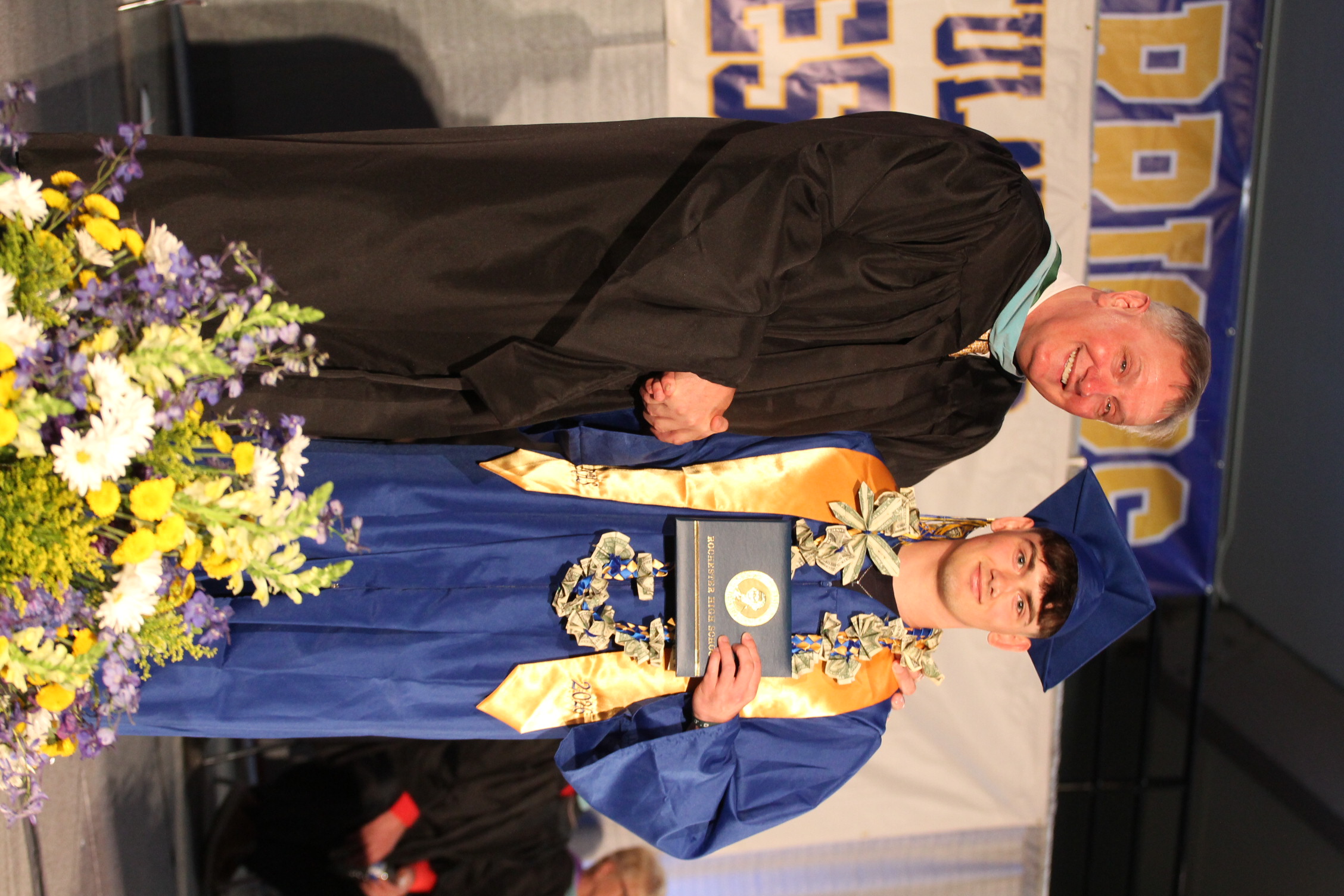 A man in a black gown and a student in a blue gown stand on a stage, holding a framed certificate, with flowers in front of them.