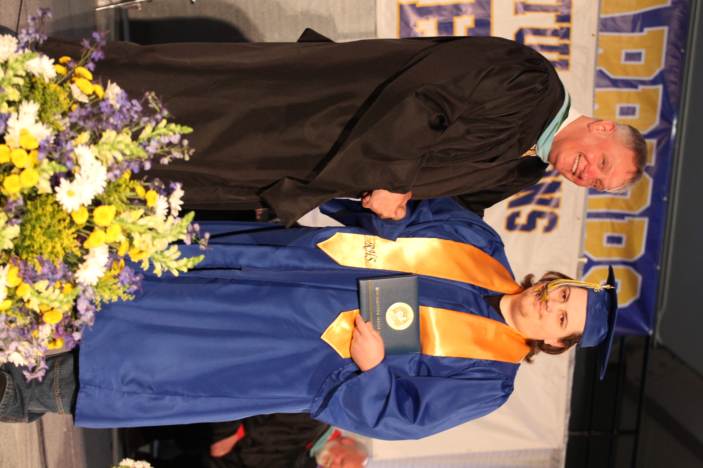 A male faculty member hands a diploma to a student. The student is on stage.