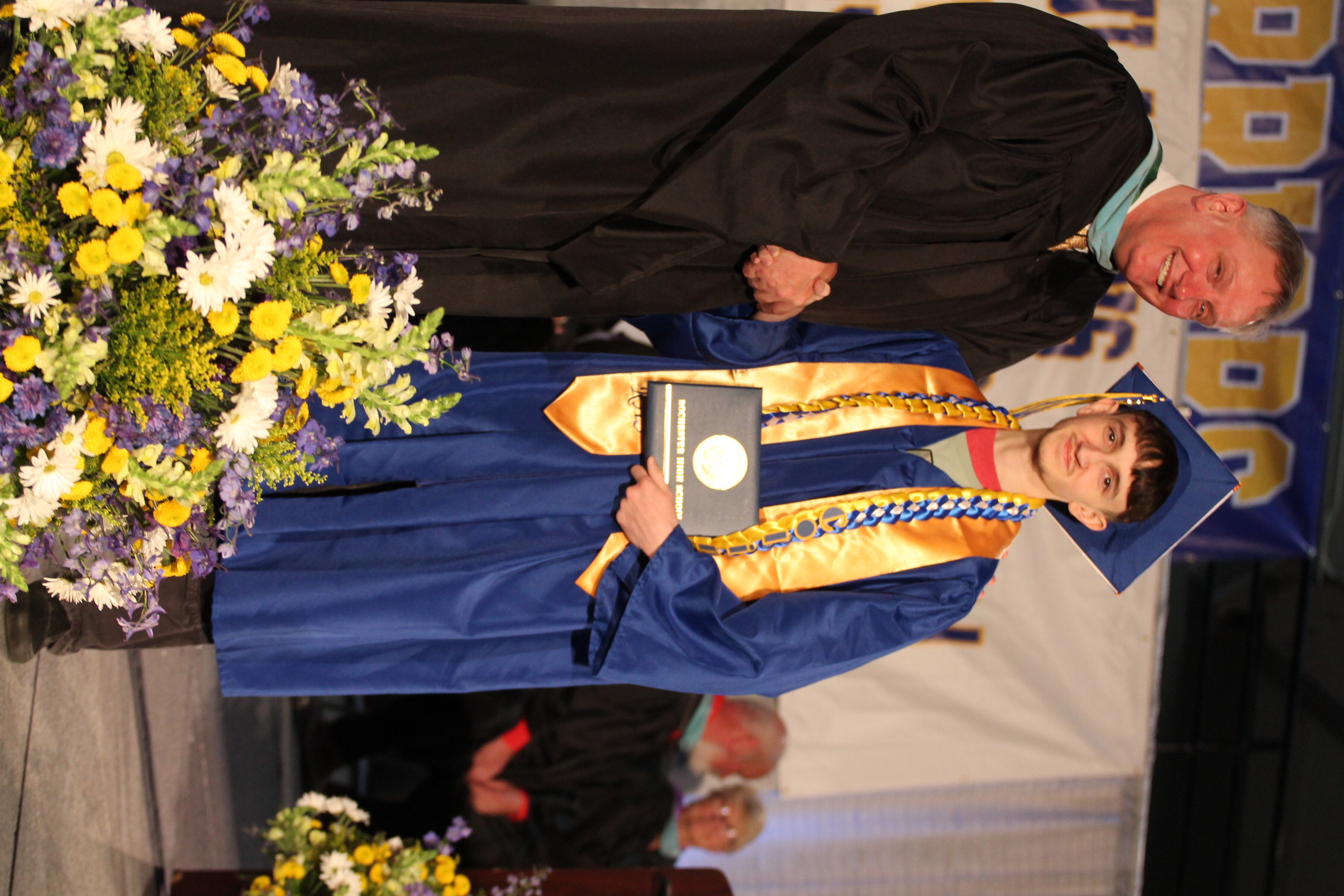 A young man in a blue gown receives an award from an adult in a black gown. They stand on stage before flowers.