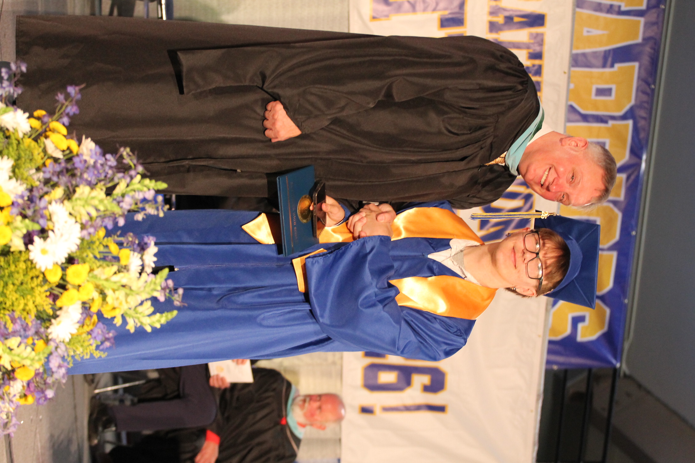A male teacher in black graduation attire shakes hands with a male student in blue. They stand on a stage with flowers.