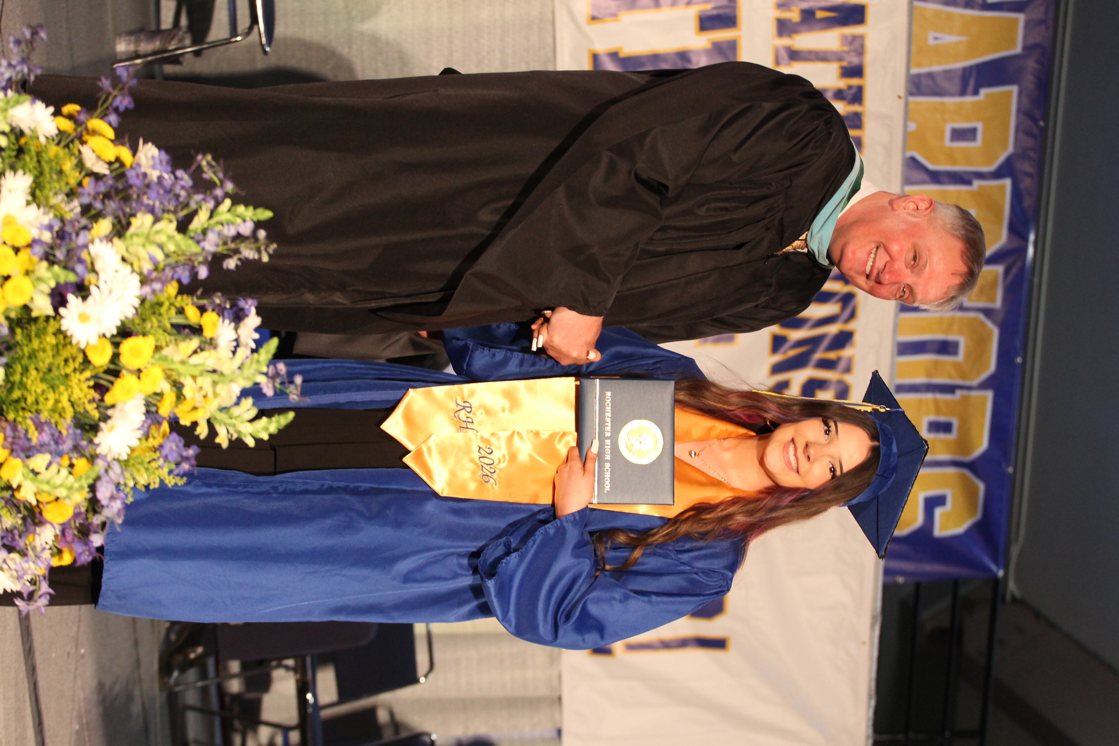 A man in a black gown hands a diploma to a woman in a blue gown. Both stand on a stage with flowers and a banner.
