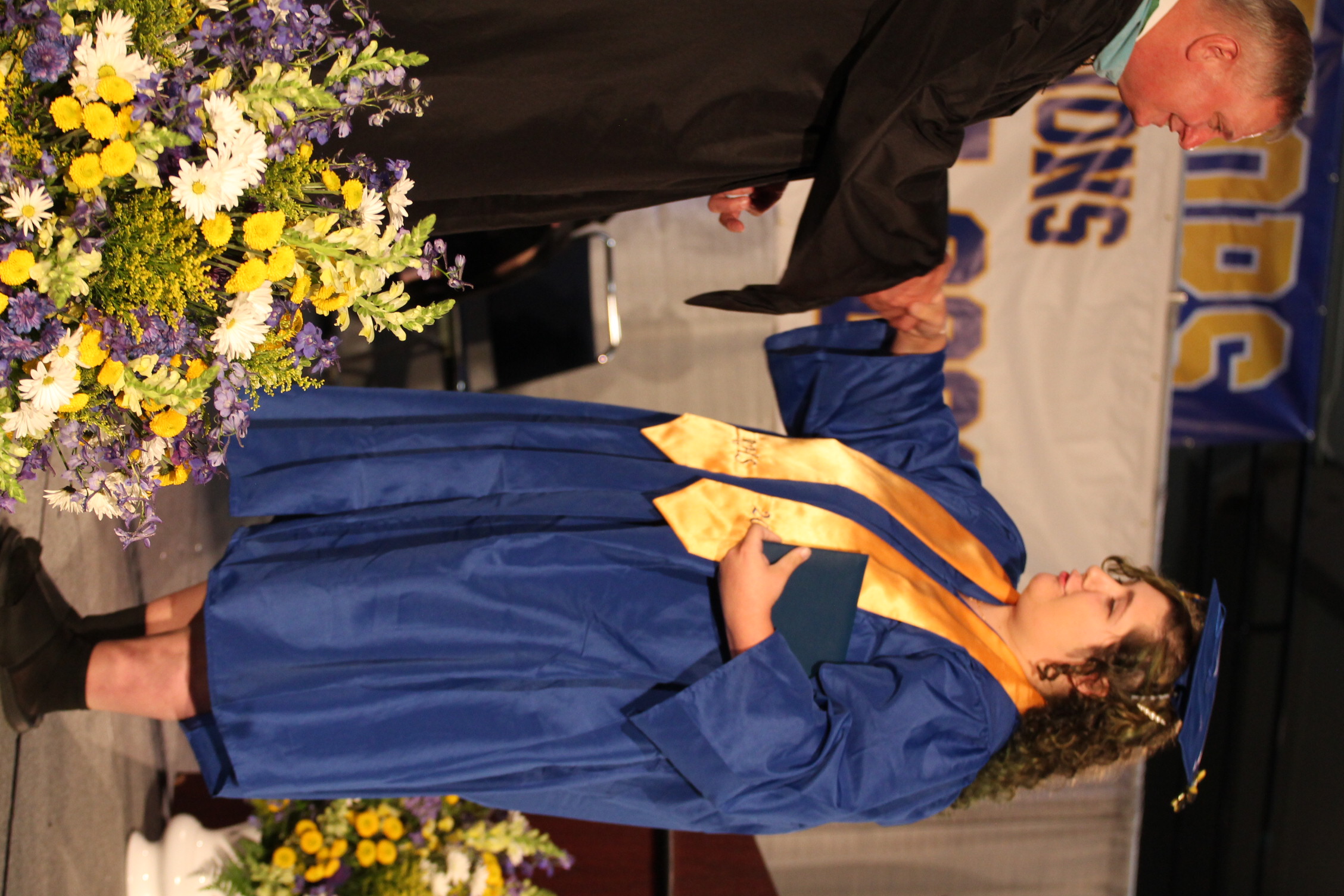 A man in a black gown hands a diploma to a woman in a blue gown. Both stand on a stage with flowers.