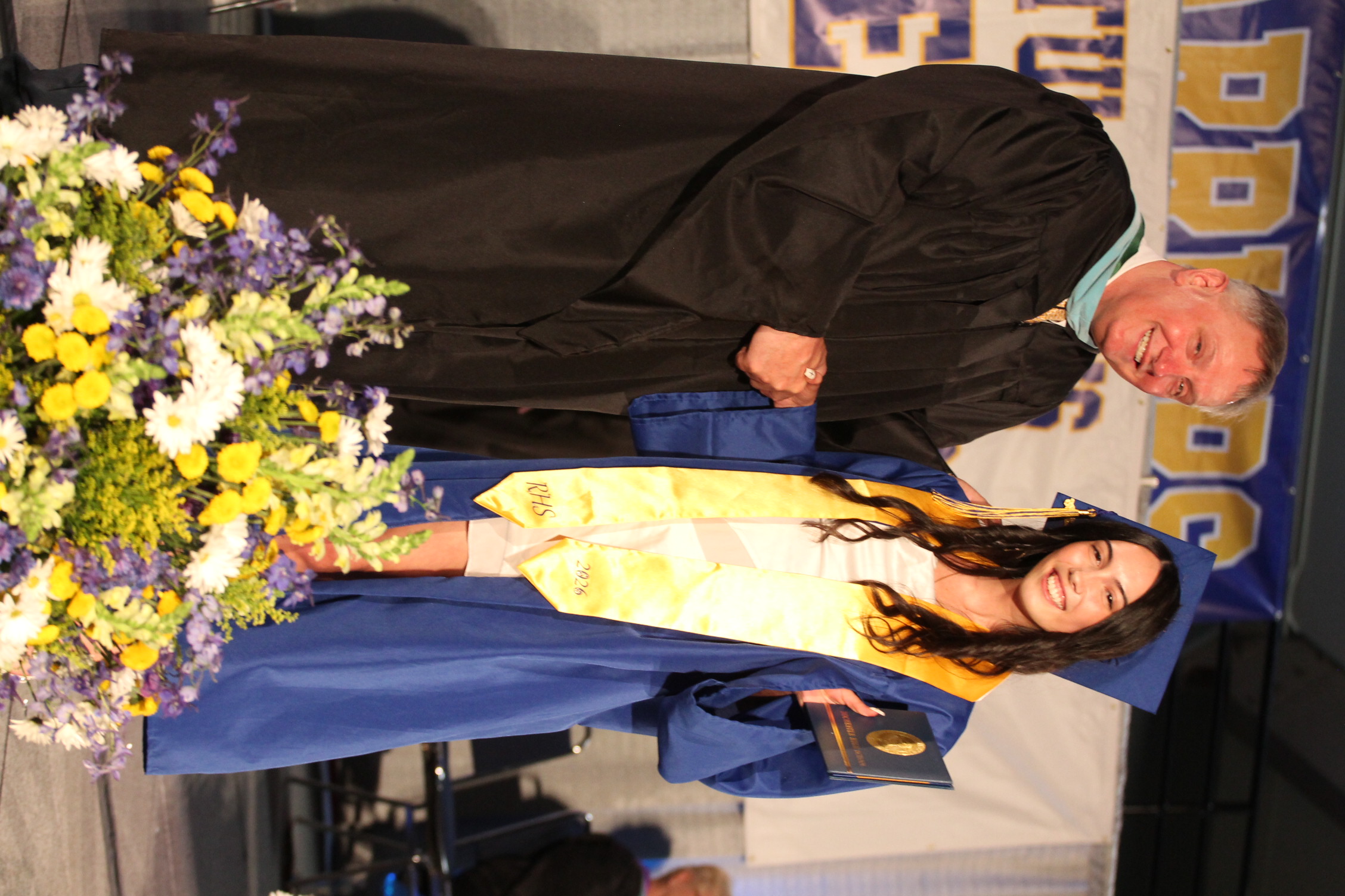 A man and woman in academic robes, smiling and shaking hands on stage with a flower arrangement.