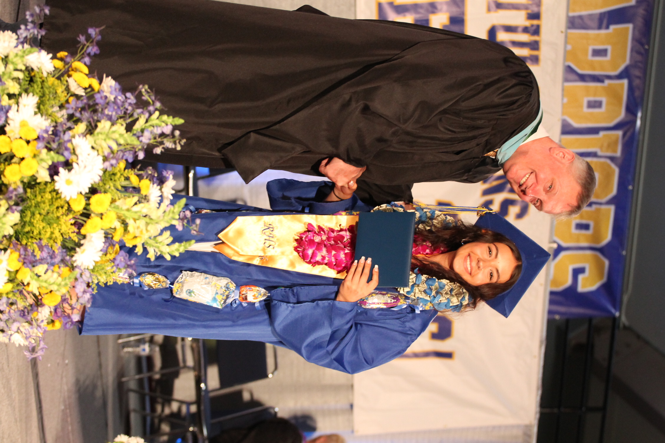A male in a black robe hands a diploma to a female graduate wearing a blue gown and a lei.