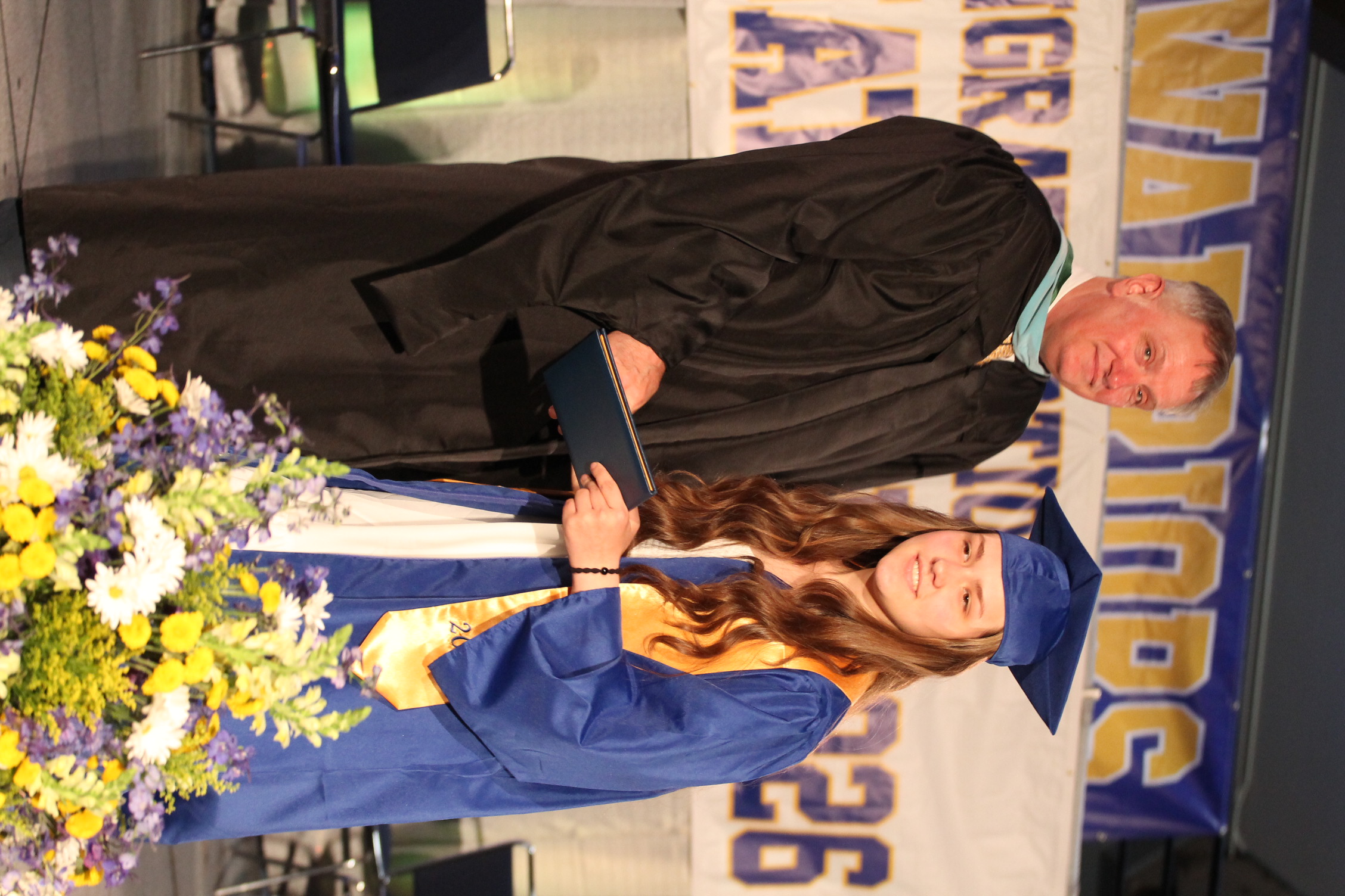 A man in a black graduation gown hands a diploma to a woman wearing a blue gown and cap.