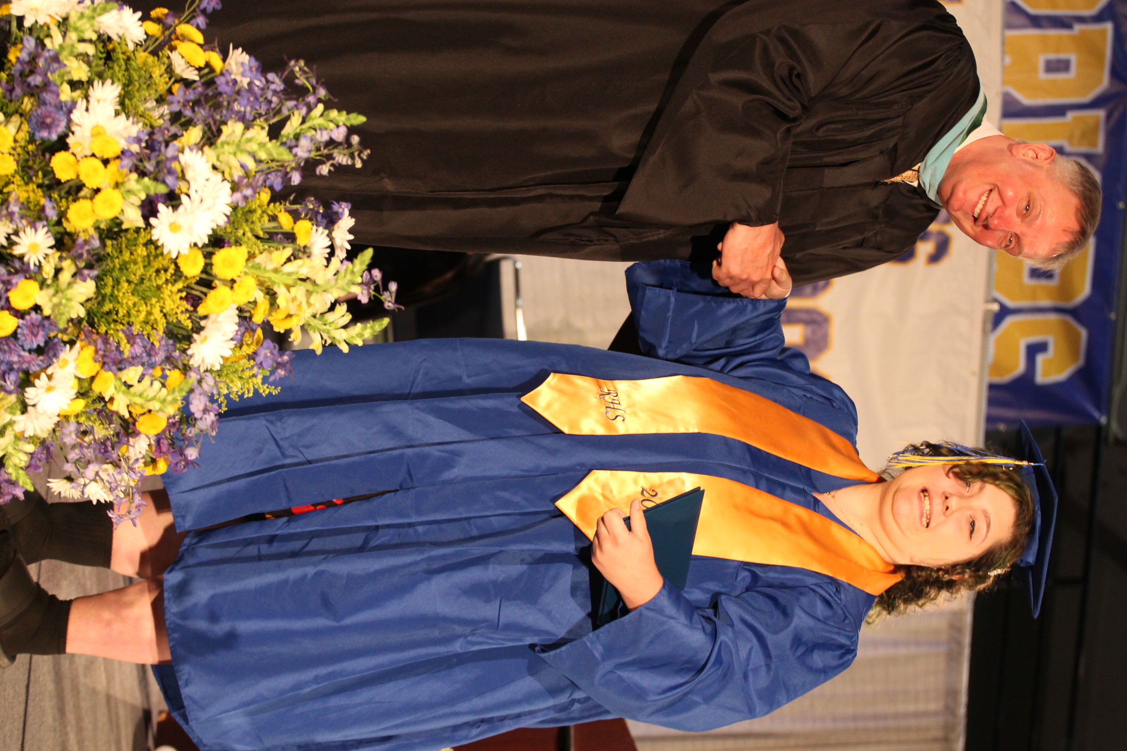 A man and woman on a stage. The man is in a black gown, the woman in a blue gown. Both are smiling, shaking hands. A flower arrangement is in front of them.