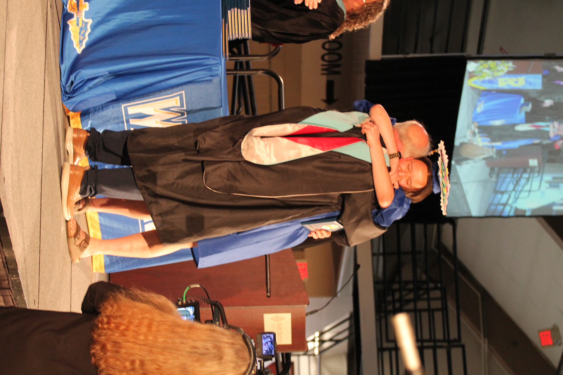 Two people on stage in academic regalia embrace. One holds a microphone, and the other wears a graduation cap.