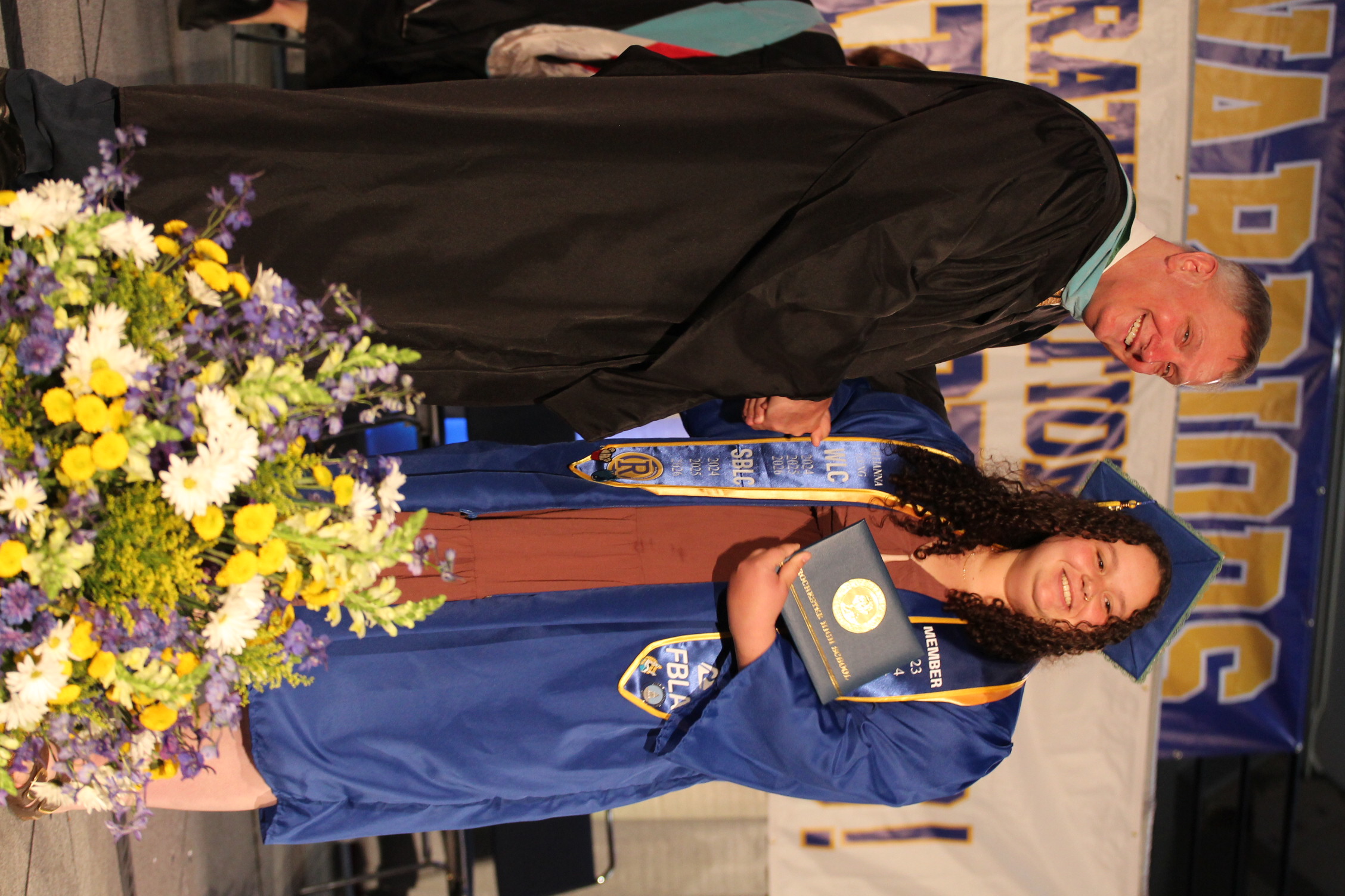 A man in a black gown hands a diploma to a woman in a blue gown. The woman holds a framed diploma and stands near a flower arrangement on stage.