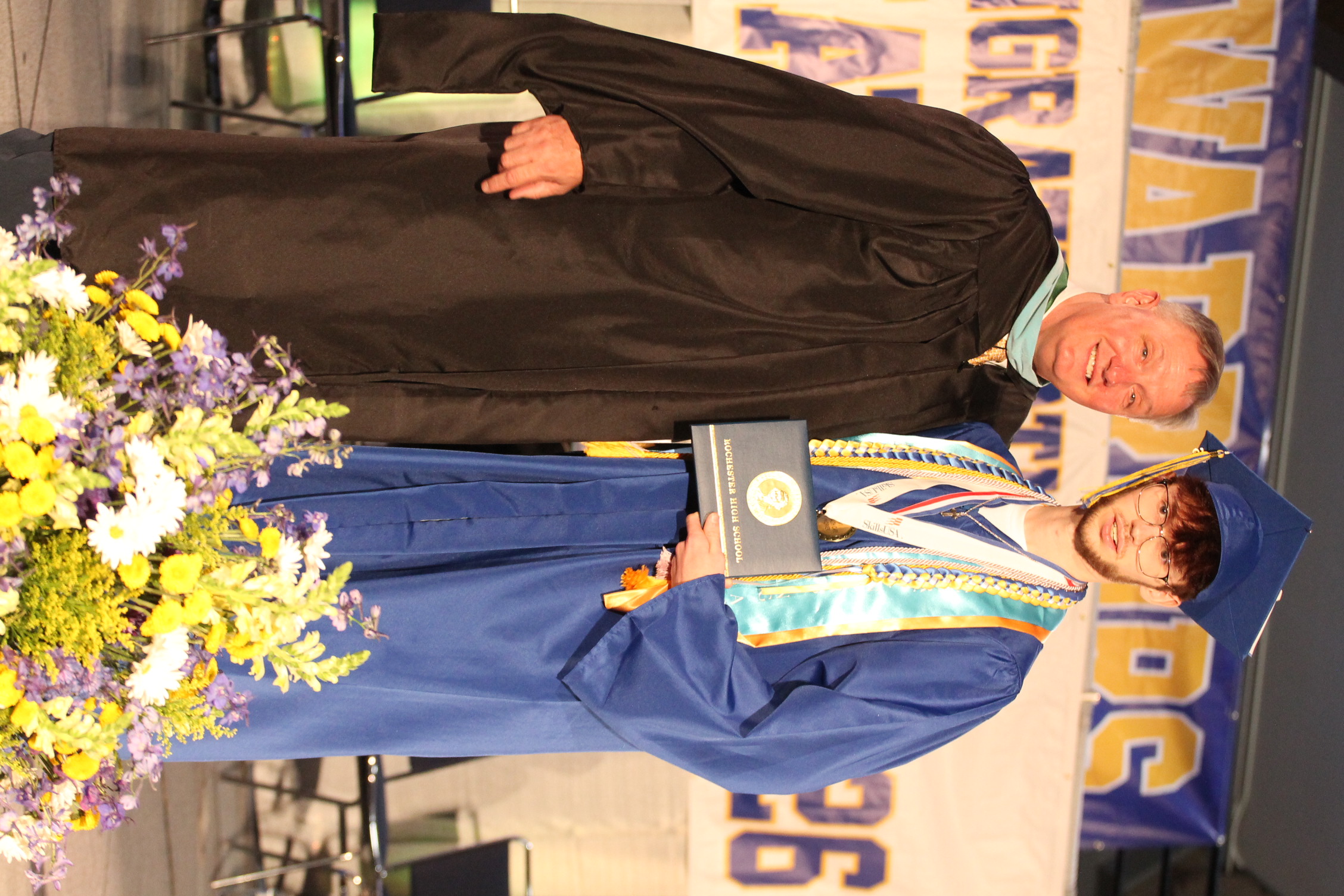 A man in a black gown and a student in a blue gown stand next to a bouquet, holding a certificate.