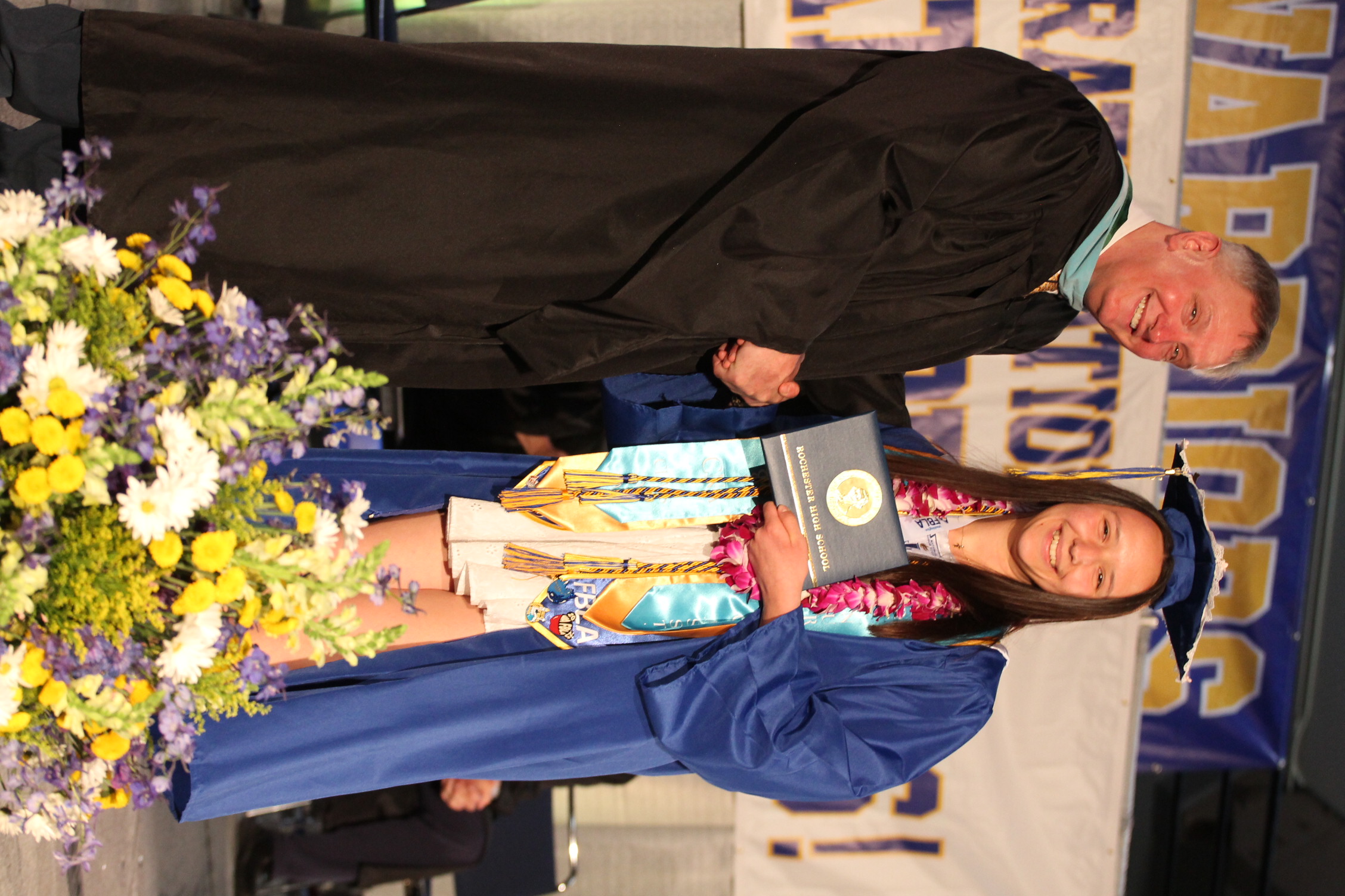 A man in a black gown shakes hands with a graduate. She wears a blue gown and holds a diploma.