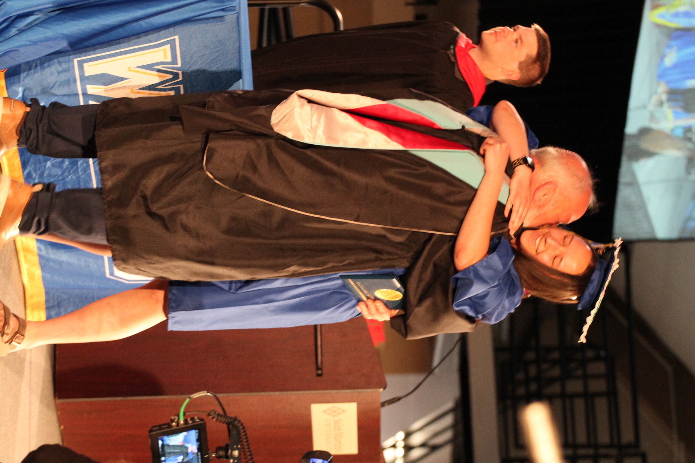 A graduate wearing a cap and gown hugs a man in front of a podium. Behind them, two people stand, one in a cap and gown.