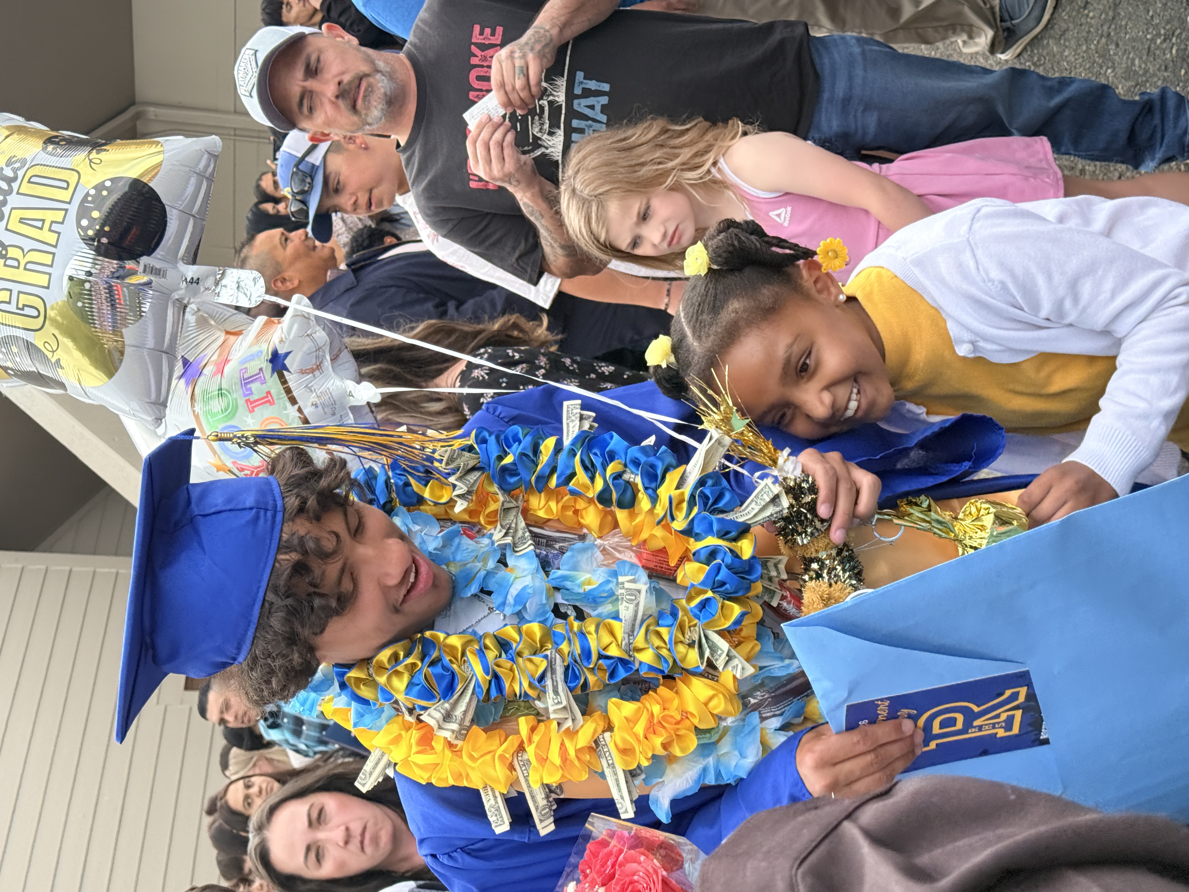 A man and a girl smile and pose for a photo. He wears a graduation cap and lei.