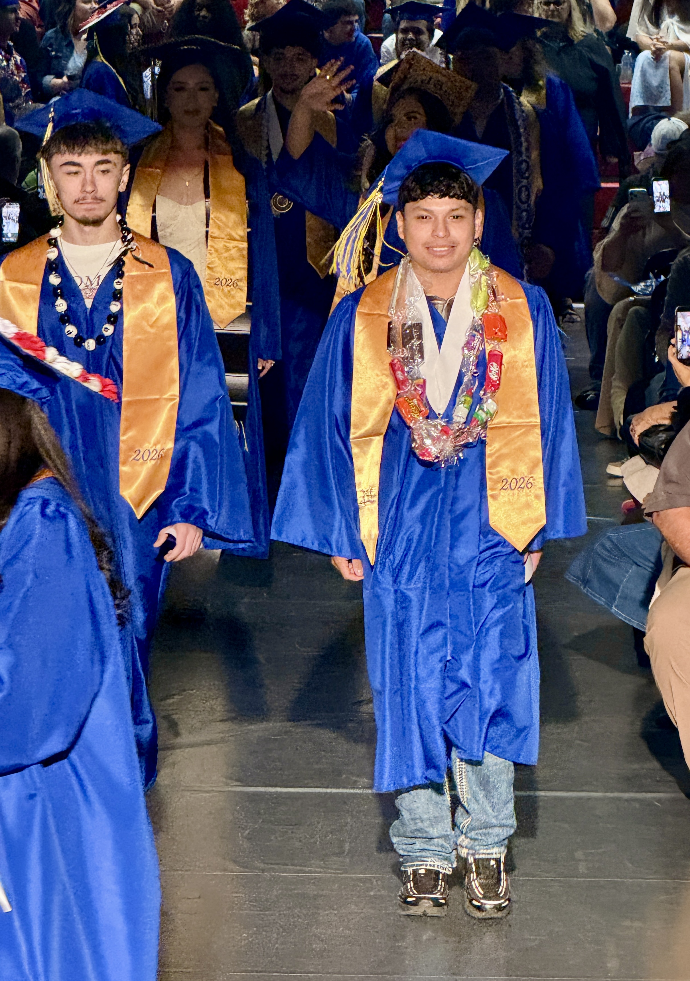 Two men in blue and gold caps and gowns walk across a stage. Behind them are seated people and some are on their phones.