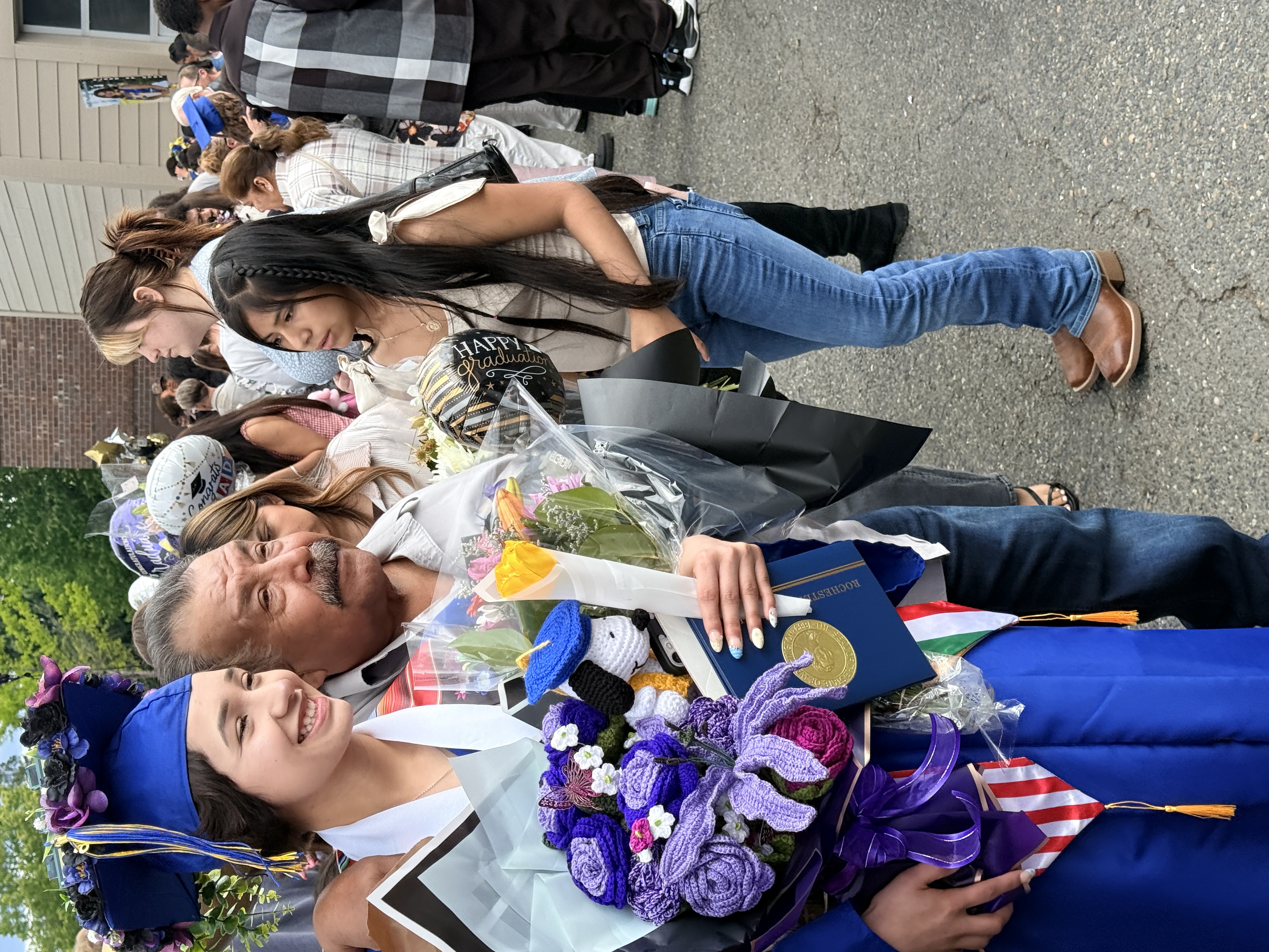 Graduates in caps and gowns hold flowers. One holds a diploma. A crowd of people is visible in the background.