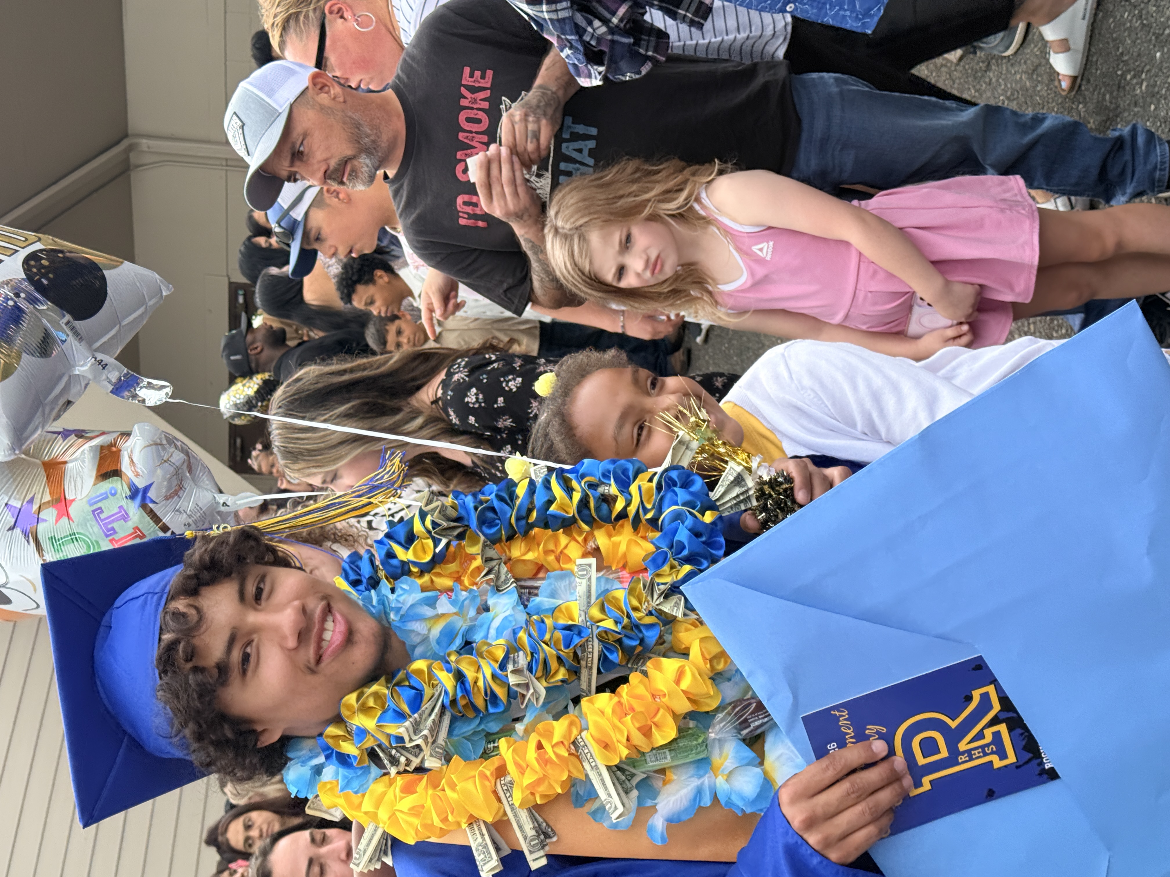 A young man in a cap and gown holds a blue bag, smiling beside two children. Behind them, people gather, some wearing hats and glasses.