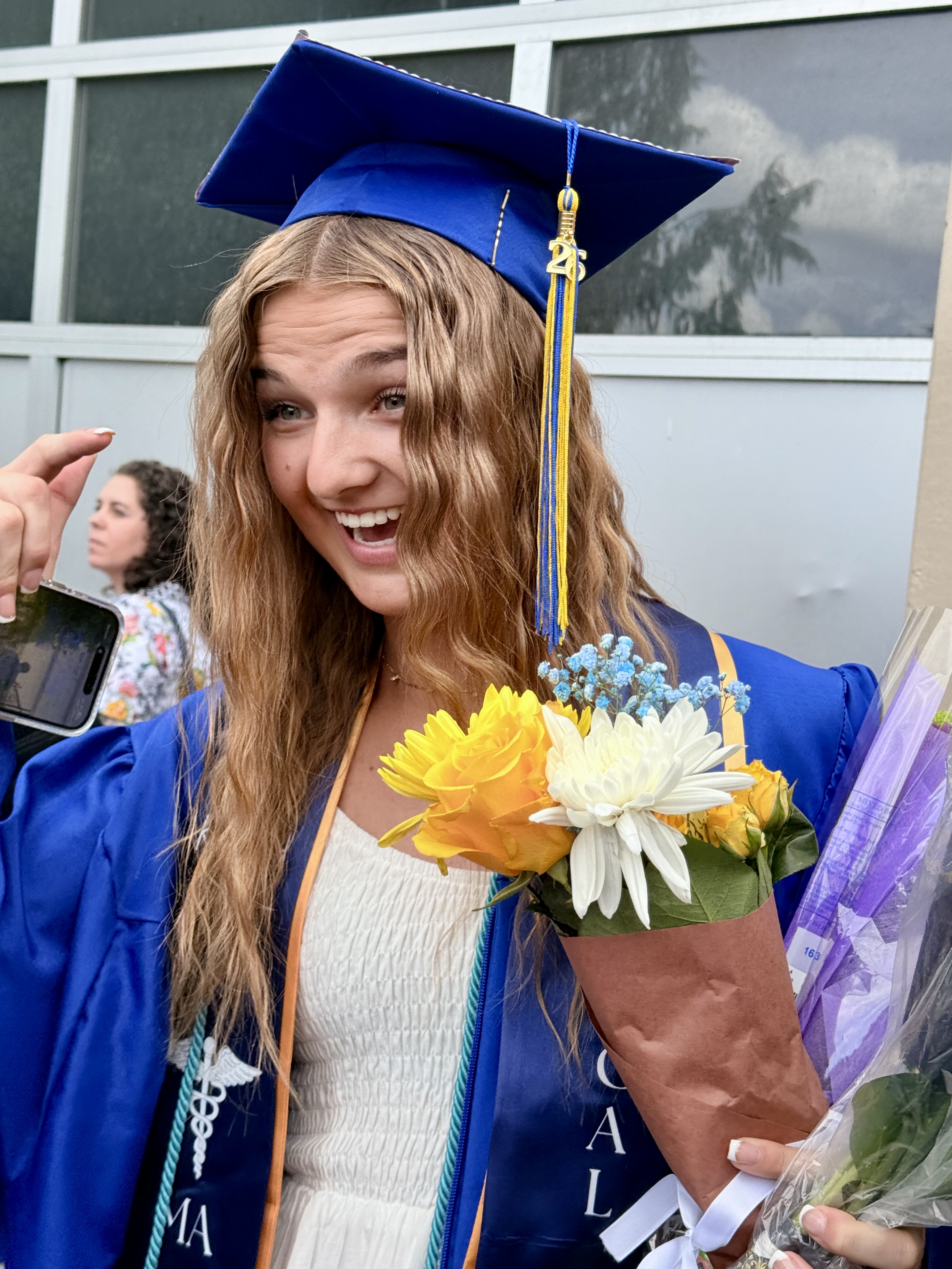 Person in blue cap and gown holds flowers and smiles, with another person in background.