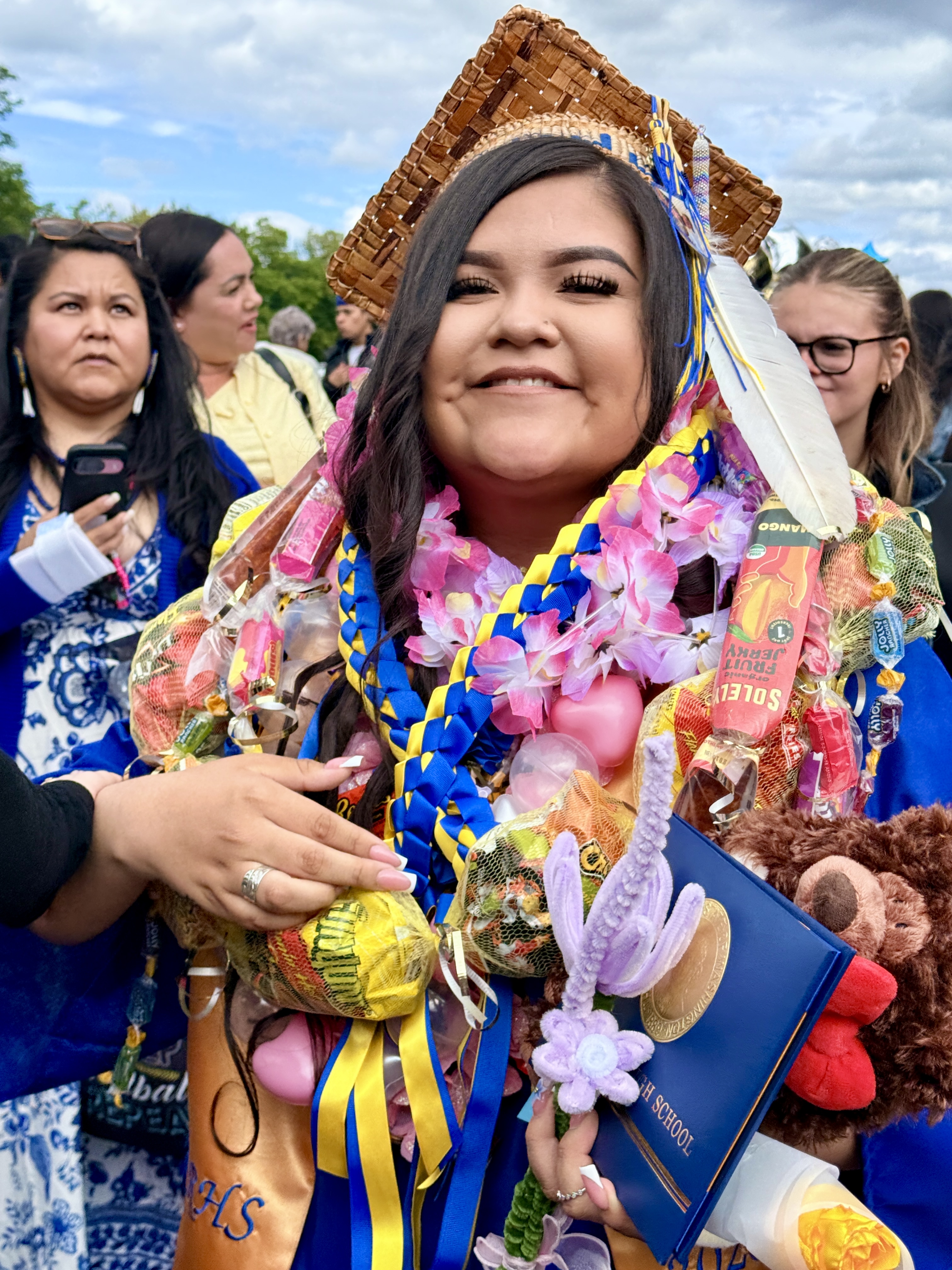 A smiling woman wearing a graduation cap, holding a book and stuffed toys, and adorned with colorful decorations.