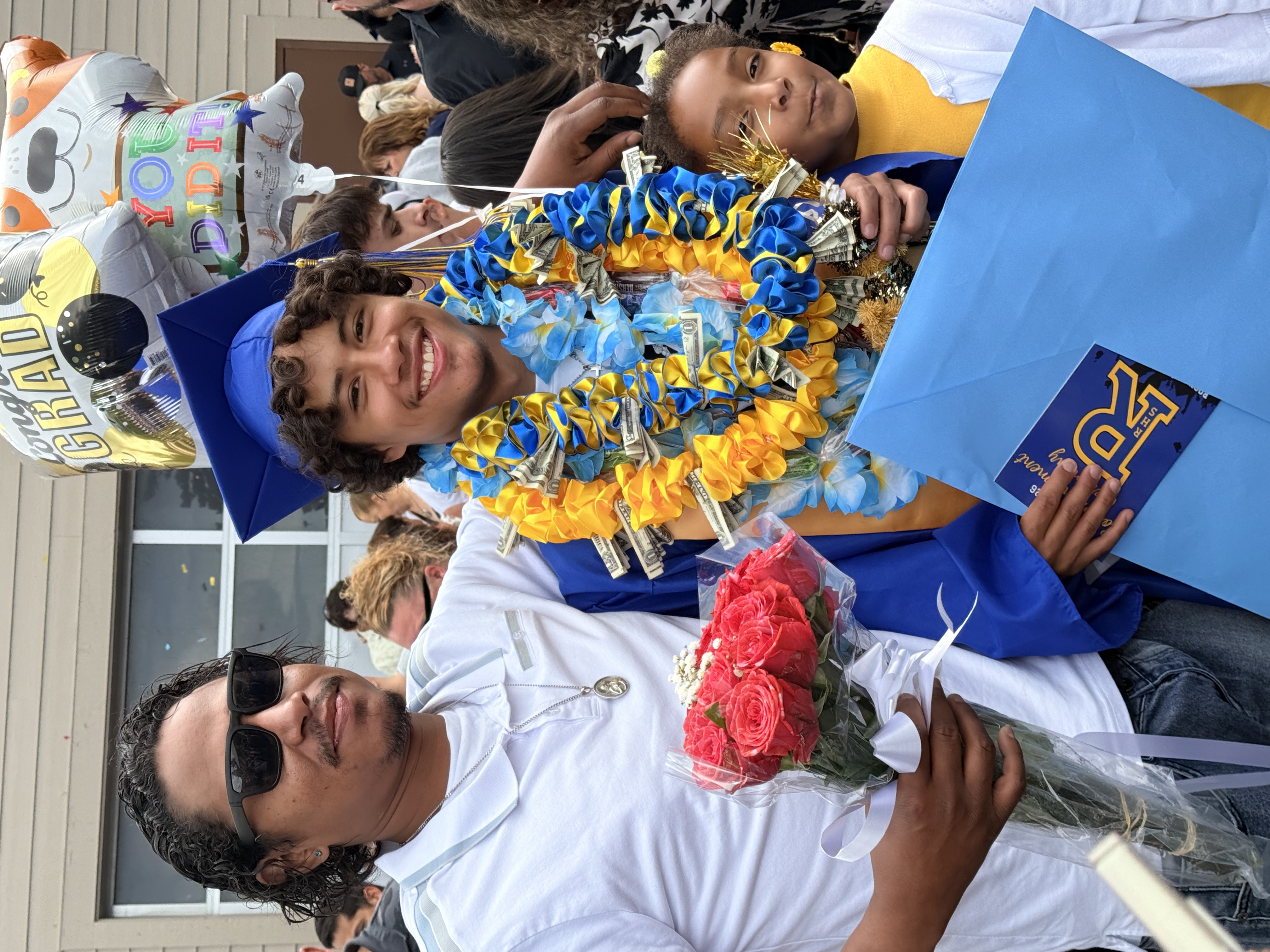 Two people smiling, one holding flowers and a cap, another with a lei and a folder.